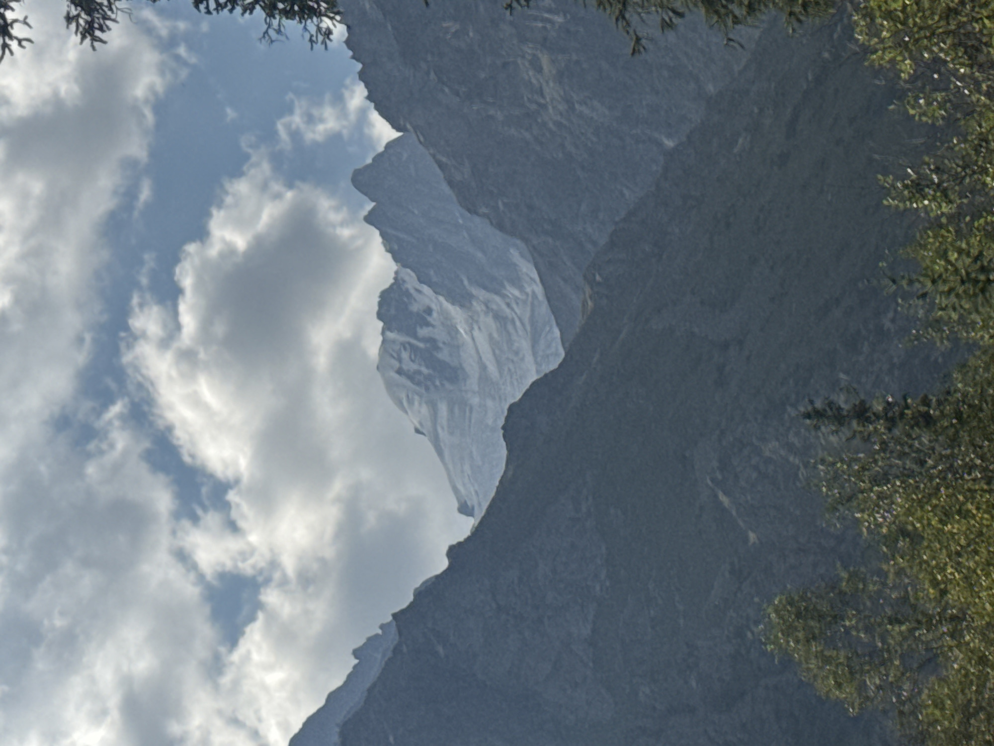 A vertical, medium-distance shot shows a wide mountain range under a cloudy sky. The foreground features a cluster of green trees, obscuring the bottom fifth of the image. Behind the trees, dark, craggy mountains rise up. In the distance, a snow-capped peak is visible, its bright white snow contrasting with the darker, rocky slopes. The sky is filled with puffy white clouds against a pale blue background. The scene is a natural landscape with no people or man-made structures visible. The location is identified as Unknown, Kyrgyzstan.