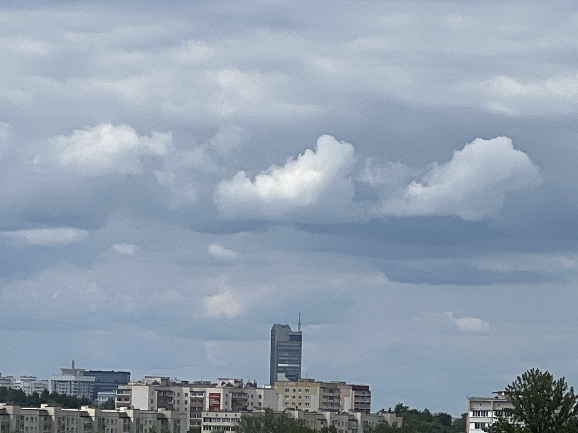 This media file depicts a cityscape under a cloudy sky. The foreground shows a dense collection of multi-story residential buildings with varied architectural styles, featuring balconies, windows, and a mix of colored facades, including some with red accents. Trees are visible interspersed between the buildings, suggesting a suburban or urban parkland setting.
Dominating the mid-ground is a tall, slender skyscraper with a modern, angular design, characterized by its glass facade and a spire-like antenna at its apex. This skyscraper stands out against the surrounding lower-rise buildings, hinting at a central business district or a prominent landmark within the city.
The sky is filled with a thick layer of grey and white clouds, indicating overcast weather. Some parts of the clouds are illuminated by sunlight, creating a contrast between lighter and darker shades. The overall lighting suggests it is daytime, possibly late morning or afternoon.
While there are no people visible in the frame, the presence of residential and commercial buildings implies an active urban environment. The scene appears to be a general view of a city, likely Minsk, Belarus, given the provided location context. There is no visible text within the image. The atmosphere conveyed is one of a typical day in a large, modern city, with a focus on the urban landscape and its prevailing weather conditions.