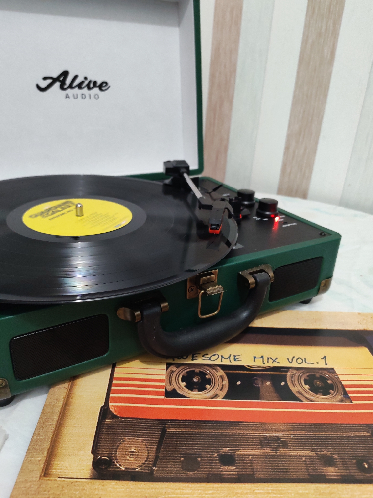 A dark green, portable record player is centrally featured, shown from a slightly elevated perspective. Its lid is open, revealing a black vinyl record with a vibrant yellow center label resting on the platter. The tonearm is positioned over the record, with the stylus engaged, suggesting the record is either playing or about to be played.
The inside of the record player's lid displays the brand name "Alive AUDIO," with "Alive" in a cursive script and "AUDIO" in capital letters beneath it. The player itself has a robust handle with brass-colored latches, and a speaker grille is visible on its side. On the control panel, black knobs are present, and a small red LED light is illuminated, indicating the device is powered on.
Beneath the record player, a decorative placemat or coaster is visible, featuring a graphic of a vintage cassette tape. The text "AWESOME MIX VOL. 1" is clearly written on the label of this cassette design.
The background consists of a light-colored, possibly striped wall or fabric, displaying vertical bands of beige, white, and a faint light-grey or pale green. The record player rests on a light-colored, possibly white, subtly patterned fabric surface, such as a tablecloth or bedsheet. The scene depicts an indoor setting, likely in daylight or under artificial light, with no people visible. No specific location cues for Izhevsk, Russia, are present.