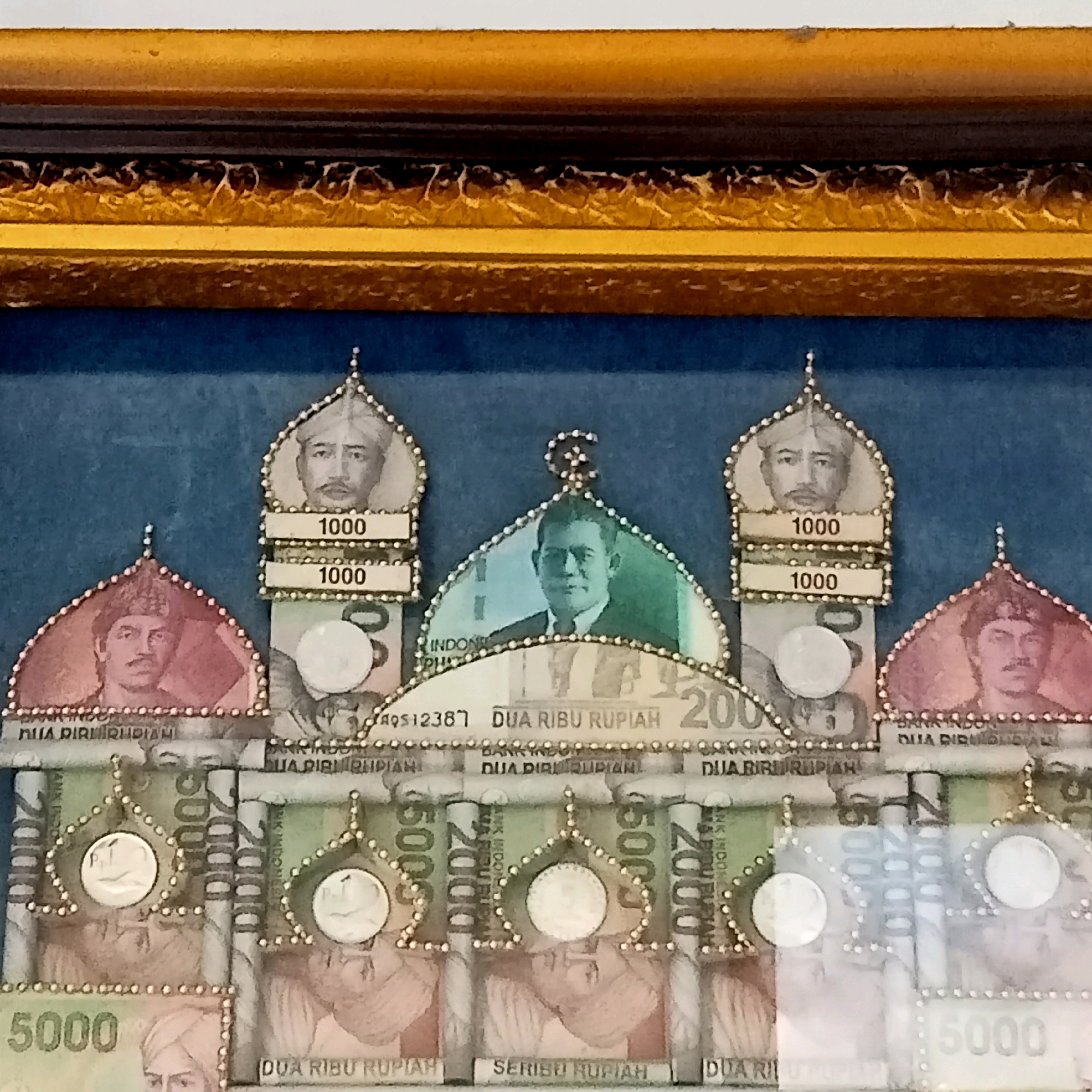 The image is a close-up of a framed artwork made from Indonesian currency. The artwork depicts a mosque-like structure, with domes and minarets fashioned from banknotes. The faces of historical figures are visible on the banknotes, along with denominations such as 1000, 2000, and 5000 Rupiah. The frame is golden and ornate. The background of the artwork is a dark blue fabric. The artwork is likely a decorative item. There is no identifiable information about the city of Jaten, Indonesia, or any specific location within it.