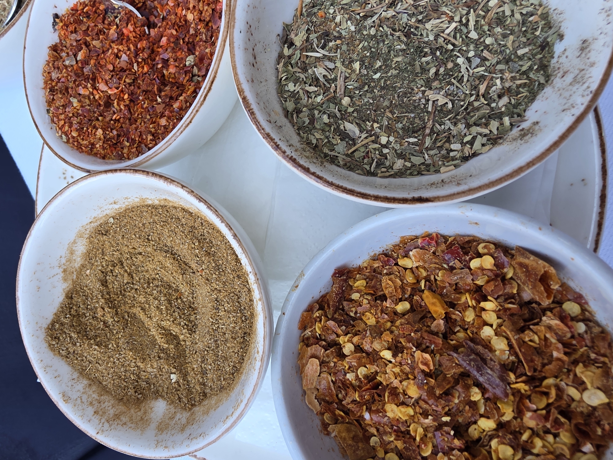 A close-up overhead shot shows four white ceramic bowls filled with various spices arranged on a white surface. From the top left, moving clockwise, the first bowl contains vibrant red chili flakes. To its right, a bowl is filled with dried green herbs, possibly oregano or a similar blend. The bottom right bowl holds more red chili flakes, this time appearing slightly larger and mixed with yellow chili seeds. Finally, the bottom left bowl contains a finely ground brown spice, likely cumin or a similar powder. The bowls have a rustic charm with a subtle brown rim. The lighting suggests it could be daytime, possibly indoors or under overcast skies. There are no people or visible text in the image. The focus is entirely on the textures and colors of the spices, implying a preparation for cooking or a display of culinary ingredients.