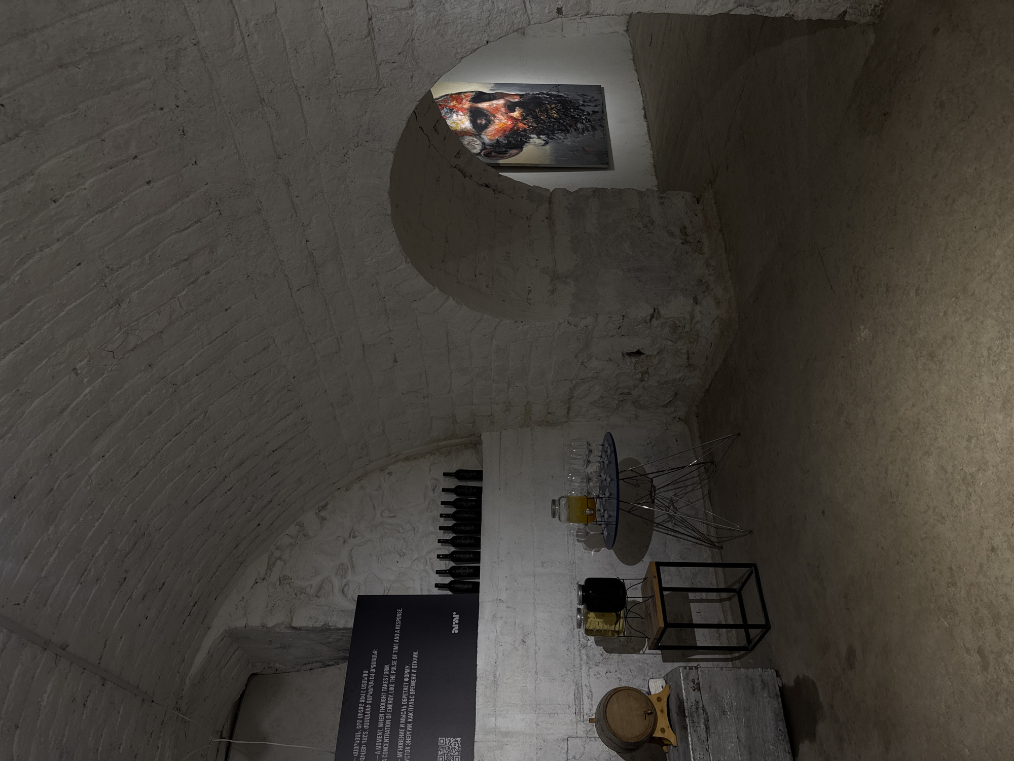 The image depicts the interior of a cellar-like space in Yerevan, Armenia, possibly an art gallery or exhibit space. The walls and ceiling are constructed of white brick, forming a curved, arched structure. A piece of artwork hangs in an alcove in the back of the room. To the left, there are wine bottles stored in the wall and a dark sign with text in Armenian and English that includes phrases like, "A moment when thought takes form." There is a small table with glasses and a jar of liquid. A black box and a small barrel are situated near a wooden box on the floor. The floor is a natural stone or concrete.