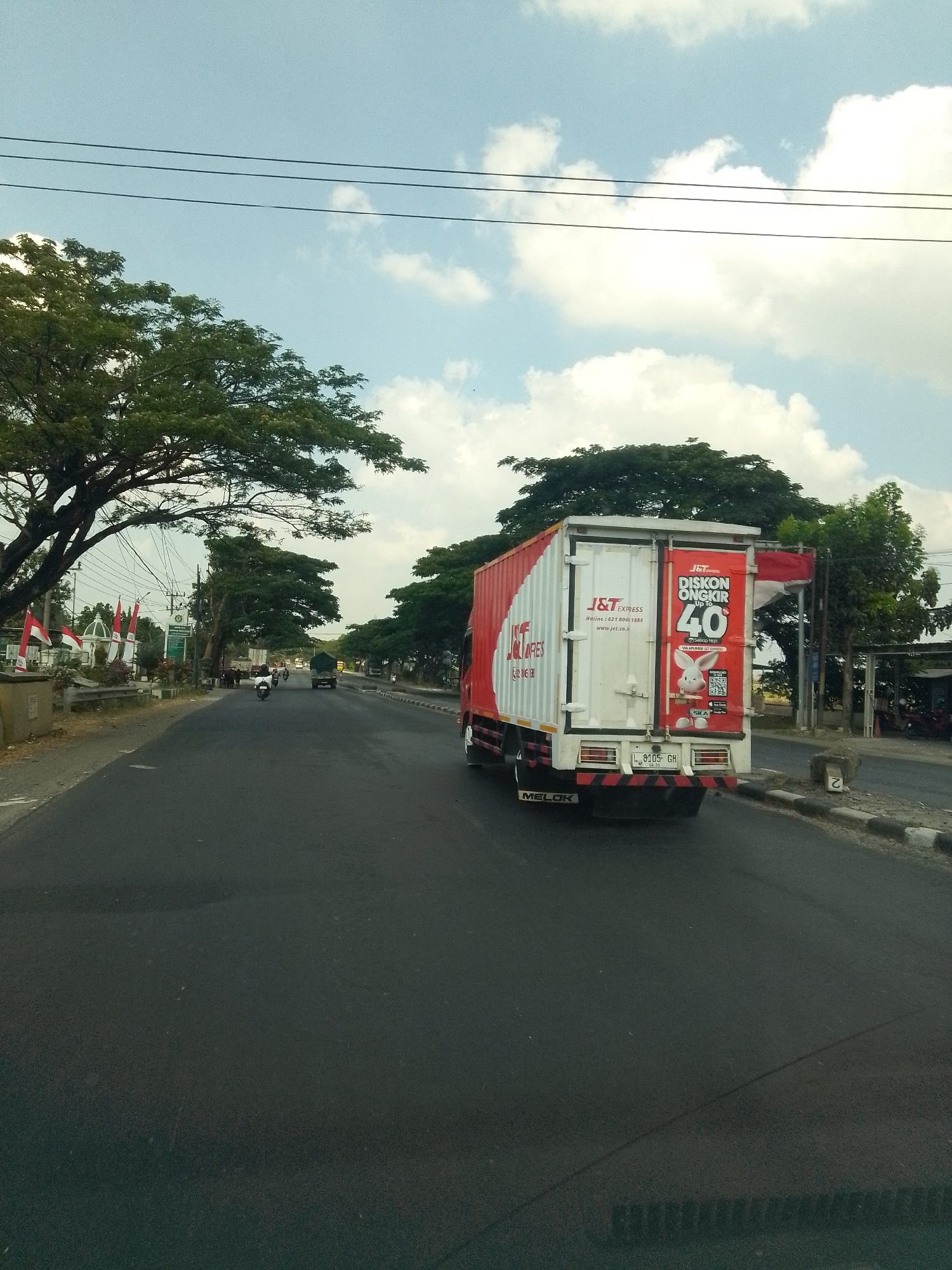 A J&T Express truck with a red and white container is driving on a paved road in Gembong, Indonesia. The truck is emblazoned with "J&T EXPRESS" and an advertisement for "DISKON ONGKIR 40". A white rabbit cartoon character is also visible on the advertisement. The truck's license plate reads "L 8105 GH". The road is lined with trees and on the left side, there are red and white Indonesian flags displayed. In the distance, other vehicles and motorcycles can be seen on the road. The sky is blue with scattered white clouds, and power lines run horizontally across the upper part of the image. The scene depicts a typical daytime highway environment in Indonesia.