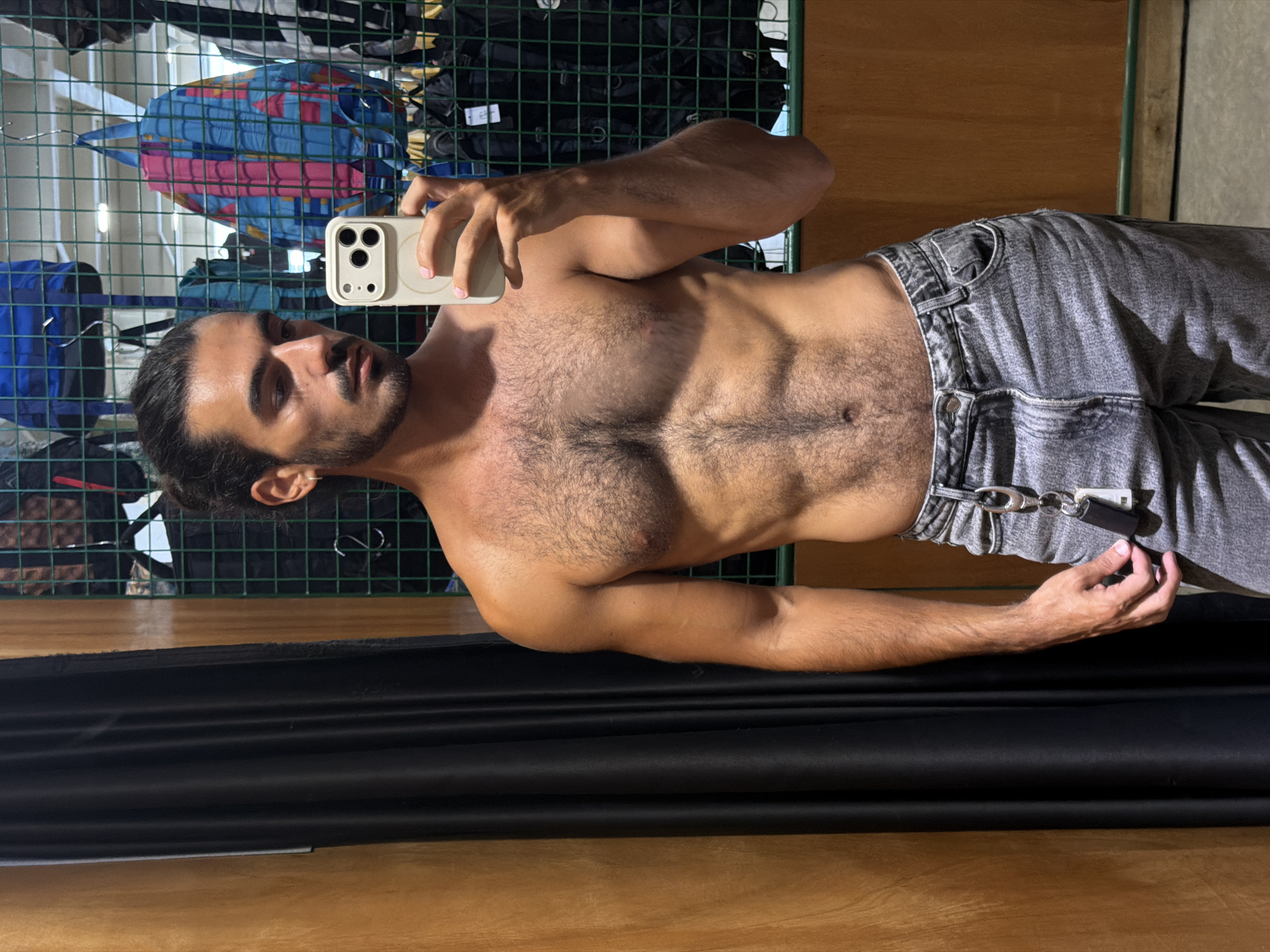 A man with dark hair pulled back, wearing grey jeans, is shirtless and taking a selfie with a phone with multiple camera lenses. The background includes a green wire grid with clothes hanging on it and wooden paneling. A black curtain is also visible in the foreground. The location is implied to be North Kuta, Indonesia, though this is not evident from the image alone.