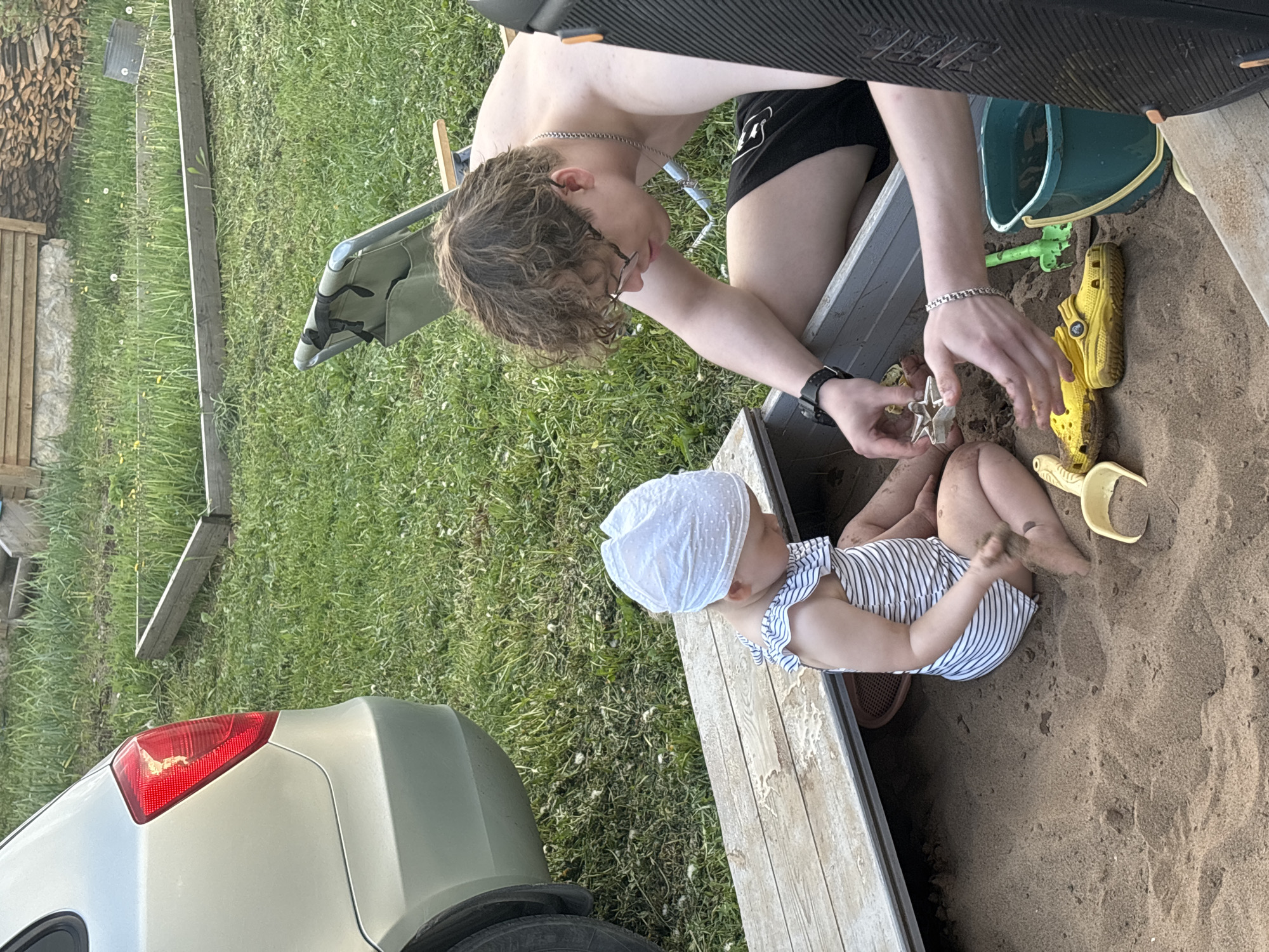 The image depicts a young child in a blue and white striped outfit and white bonnet sitting in a sandbox. An older person, shirtless with curly brown hair and glasses, is kneeling next to the sandbox, engaging with the child. In the sandbox are several yellow plastic toys including a shoe, a bucket, and a shovel. A car is visible on the edge of the sandbox. The setting is outdoors, in a grassy area. The location is Пальники, Russia.