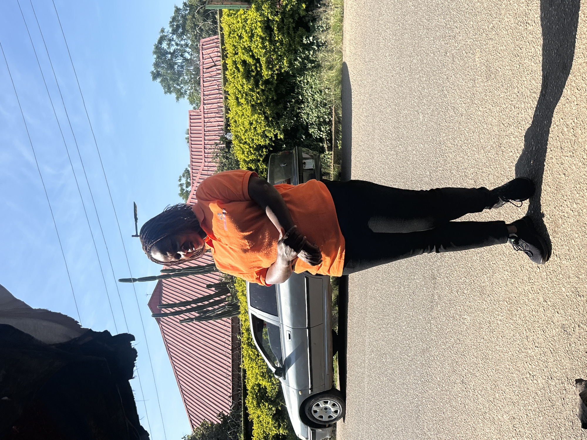 A full-body, medium shot captures a woman standing on an asphalt road in Jos, Nigeria. She is facing the camera, with her body angled slightly to the left. The woman is wearing an orange polo shirt and black pants, complemented by black athletic shoes. Her arms are crossed in front of her, and her hair is styled in braids. Behind her, a silver sedan is parked, and beyond that, a vibrant green hedge runs along a property line. A red-roofed building with white trim is visible behind the hedge, partially obscured by several tall, spiky plants. The sky above is clear blue, with a few thin clouds. The scene is illuminated by bright sunlight, casting a distinct shadow of the woman on the road to her right. The foreground shows a portion of the asphalt road and a dark, out-of-focus object, possibly an animal, on the lower left side of the image.