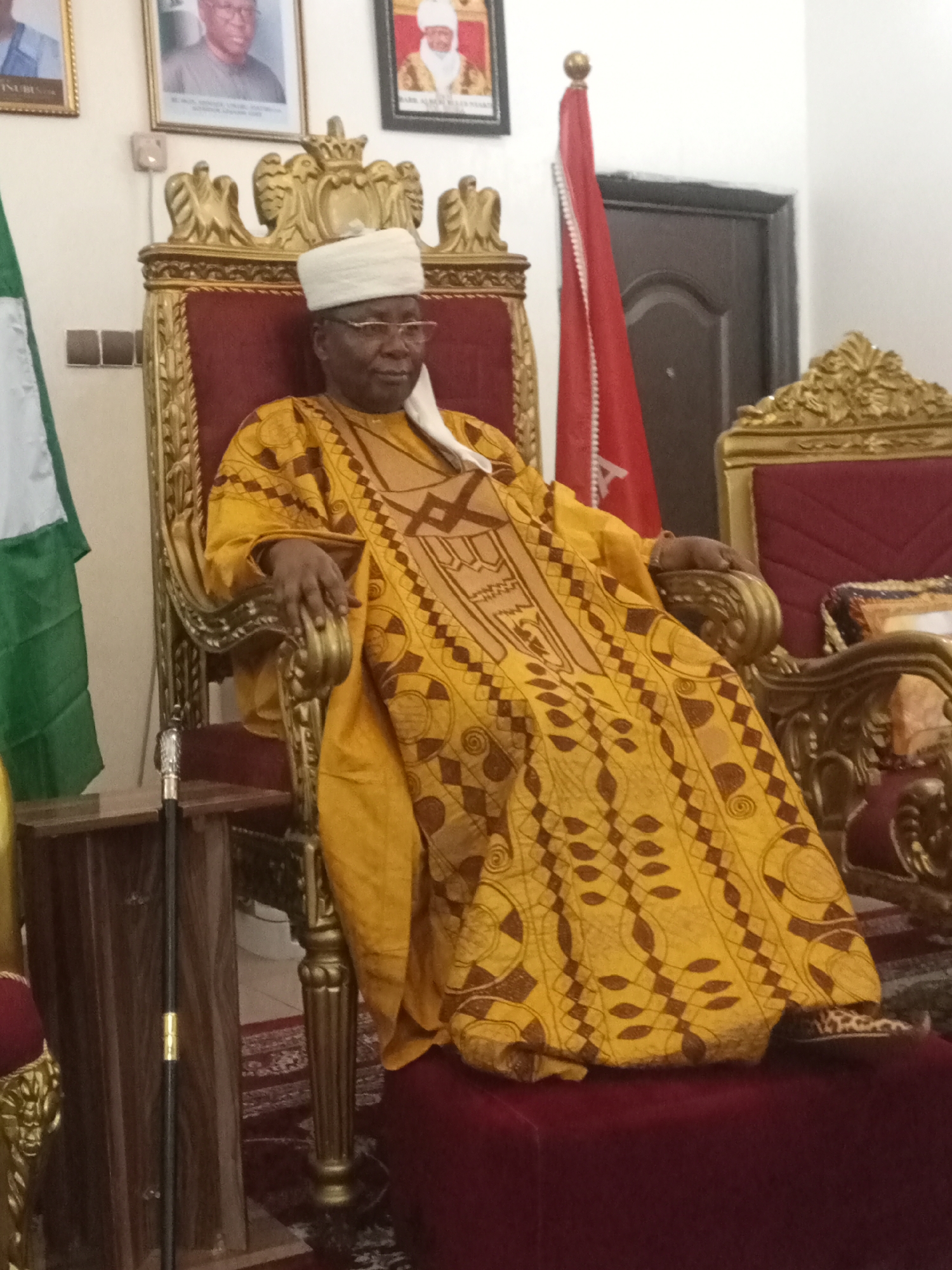 In an indoor setting, an elderly man sits on an ornate golden chair with a red cushion. He is dressed in a yellow robe with brown geometric patterns and a white cap. He wears glasses and has a fair complexion. To his left, there is a Nigerian flag, and a walking cane stands nearby. Behind him, there are two framed portraits hanging on the wall. To his right, another ornate chair is visible, partially obscured. A red flag is also present in the background, along with a dark-colored door. The location is the city of Unknown, Unknown.