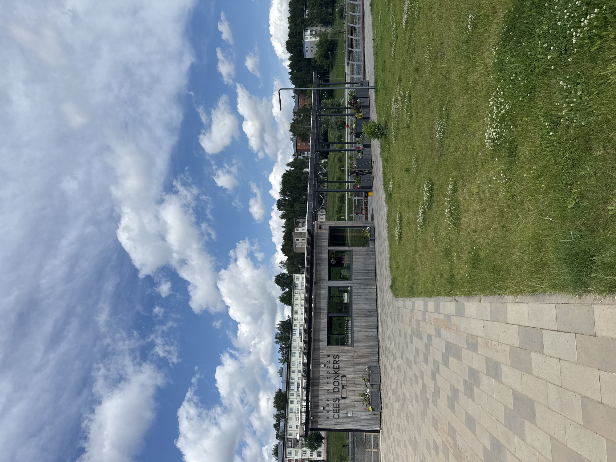 The image shows a modern wooden cafe building with the text "CAFE DONKERS" and glasses logo on its facade, located in the city of Votkinsk, Russia. The cafe is situated on a paved walkway next to a grassy area with scattered white wildflowers. In the background, there are trees, a body of water, and several apartment buildings. The sky is blue with scattered white clouds. The scene appears to be a sunny day, with no people visible in the immediate vicinity of the cafe or walkway. The cafe has large windows, and some plants are visible in planters outside the building and under a covered seating area.