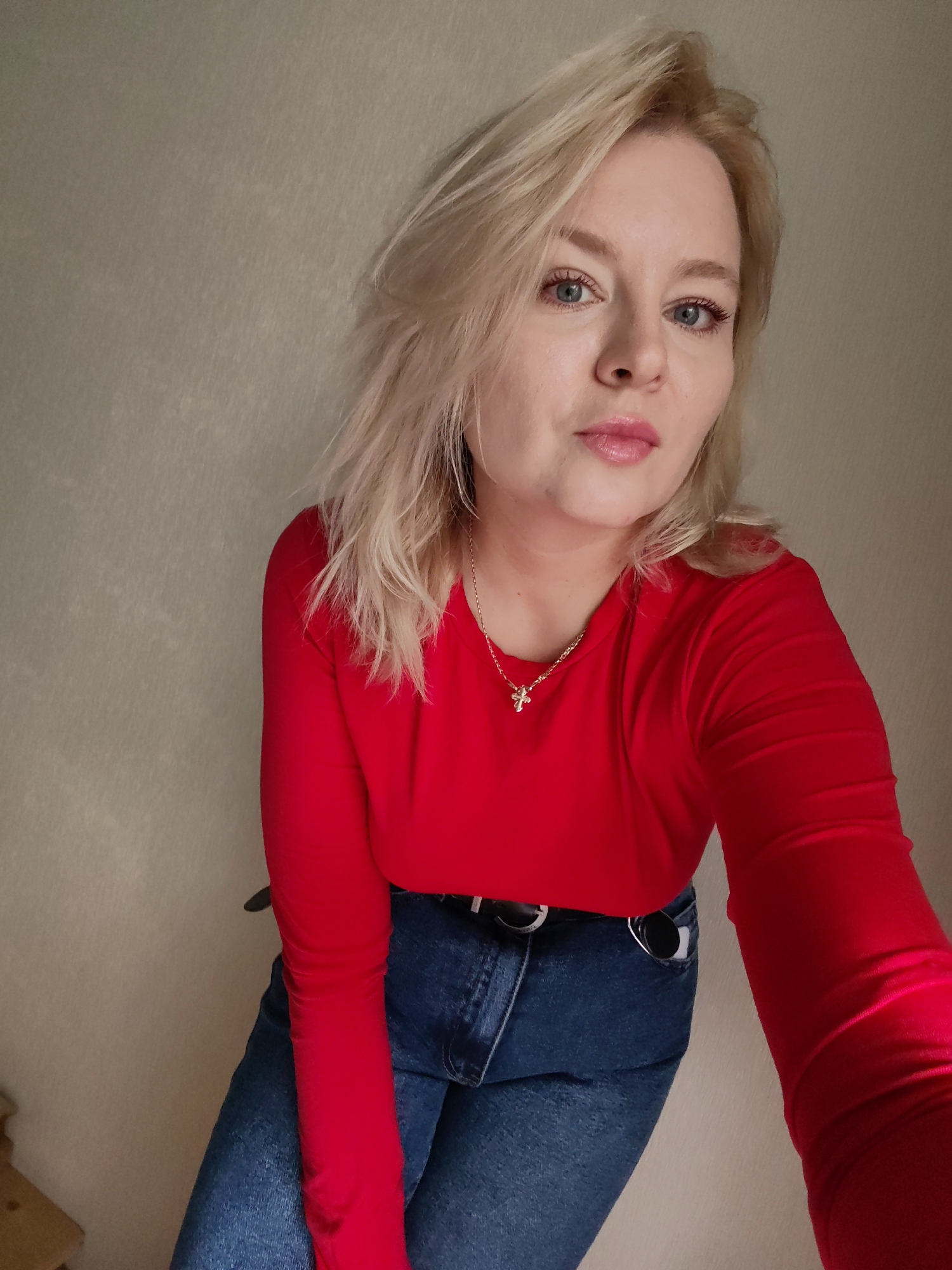 The image shows a woman with blonde hair and blue eyes, wearing a red long-sleeved shirt and blue jeans. She is looking at the camera with a neutral expression. The background is a plain, light-colored wall with some textured shadows. The lighting is soft and diffused. The scene appears to be indoors, possibly in a home or studio. The image is a selfie, as indicated by the angle and the fact that the woman's arm is visible extending towards the camera. There are no specific landmarks or identifying features that would allow for pinpointing a location within Izhevsk, Russia.