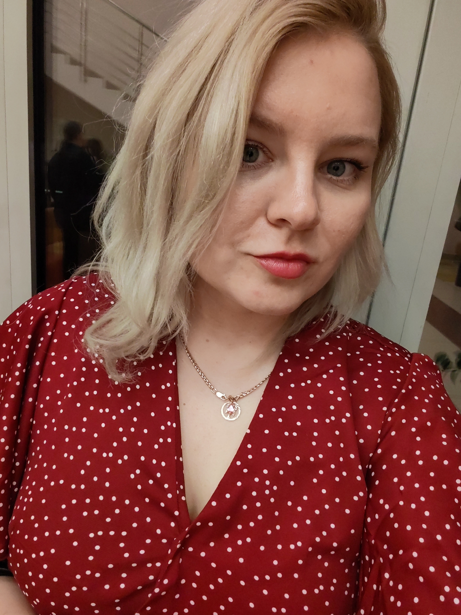 A medium close-up, head-and-shoulders portrait captures a woman indoors, likely a selfie. The woman has fair skin, light blonde, wavy hair that falls around her shoulders, and blue-grey eyes. She wears a red dress adorned with white polka dots and a V-neckline. Around her neck is a gold-toned chain necklace with a circular pendant featuring an intricate design, possibly a stylized tree, and a small pink or rose-colored gem. She has light makeup, including pinkish-red lipstick and eyeliner, and looks directly at the camera with a neutral to slightly alluring expression.
In the blurred background to her left, a reflection in a glass surface reveals a modern indoor setting with a staircase and two dark, indistinct human figures. The lighting in the image suggests artificial indoor illumination. No text is visible in the image. The setting is in Izhevsk, Russia.
