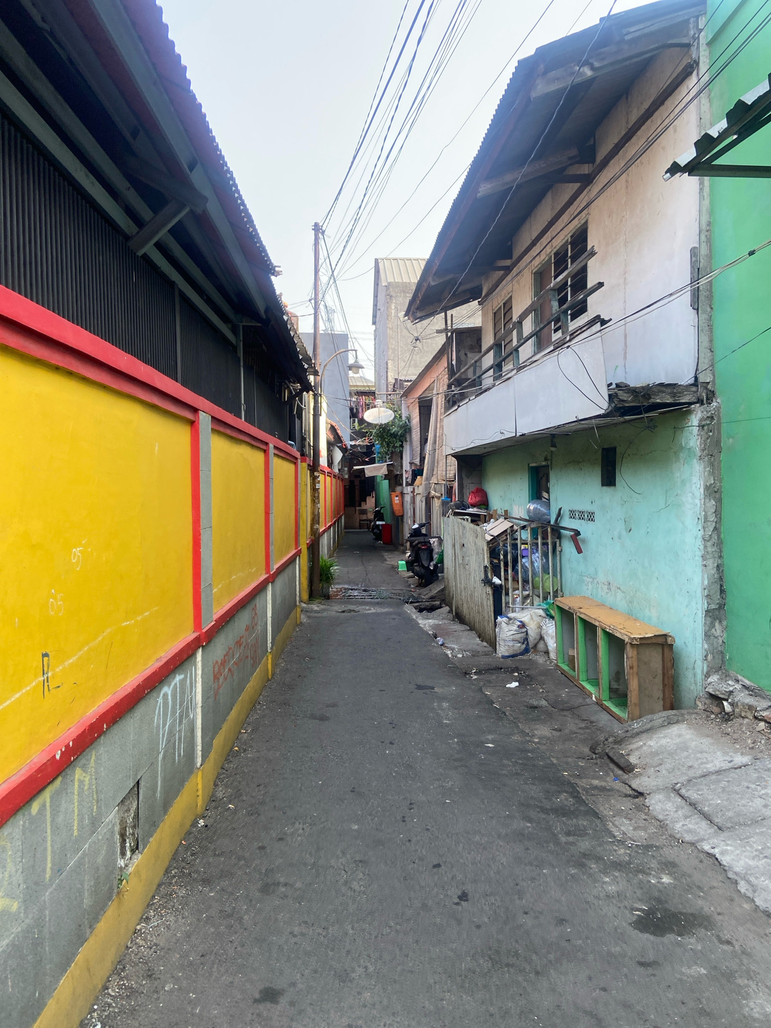 The image depicts a narrow alleyway in North Jakarta, Indonesia, flanked by buildings on either side. On the left, a tall, grey corrugated metal wall is painted with a bright yellow lower section, bordered by red trim. Several faint markings, possibly numbers, are visible on the yellow wall. The right side of the alley features a light blue building with a weathered appearance and a green upper section. The building has multiple windows and a precarious-looking wooden balcony. Items are stored in front of the blue building, including sacks of material, a wooden shelf unit, and a motorcycle parked further down the alley. Electrical wires crisscross overhead. The alley pavement is asphalt and appears somewhat worn, with patches of darker and lighter color. Trash is scattered along the sides of the alley. The overall impression is one of a densely populated urban environment with signs of wear and tear.