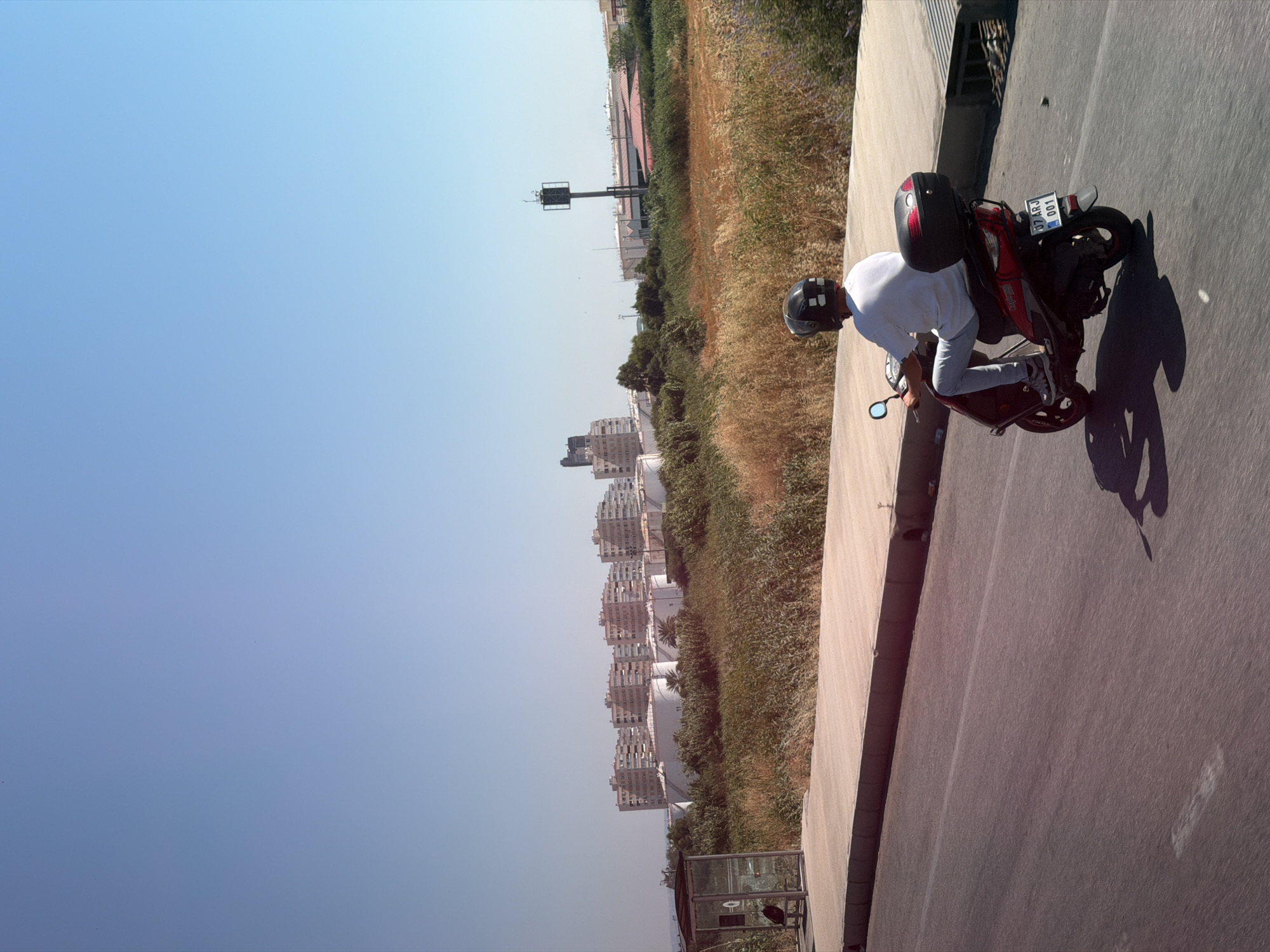 In the city of Konyaaltı, Turkey, under a clear blue sky, a person wearing a white t-shirt and light-colored pants rides a red scooter. The rider has on a black helmet. The scooter has a black storage box on the back, and a license plate that reads "07 ARJ 001". They are on a paved road, with a concrete sidewalk to their right. Beyond the sidewalk is a patch of dry, overgrown grass and vegetation. In the background, there are several large, white cylindrical tanks, and behind them, a row of apartment buildings. Further in the distance, a tall structure with a square metal frame at the top stands on a pole. A glass bus shelter with a bench inside is visible to the far left of the image, next to the sidewalk.
