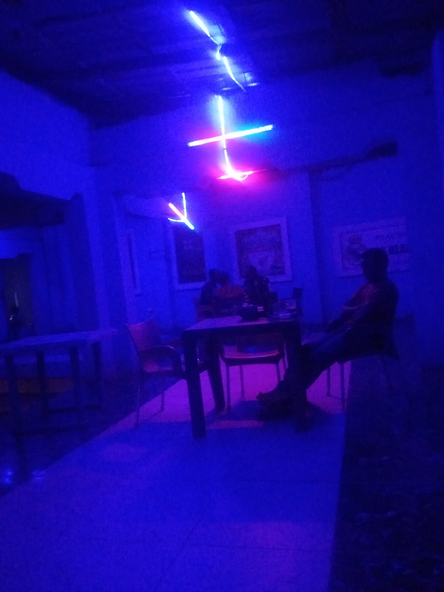 The image depicts an interior scene illuminated by blue and pink neon lights, suggesting a bar or lounge environment in Sabon Gari, Nassarawa, Nigeria. Two individuals are present. One, a man, is seated on a chair at a table, his body angled away from the camera. He appears to be wearing a patterned shirt and dark pants. The other individual is partially visible, also seated at the table. The room contains several tables and chairs, with framed artwork on the walls. The overall ambiance is dim and atmospheric, due to the colored lighting.