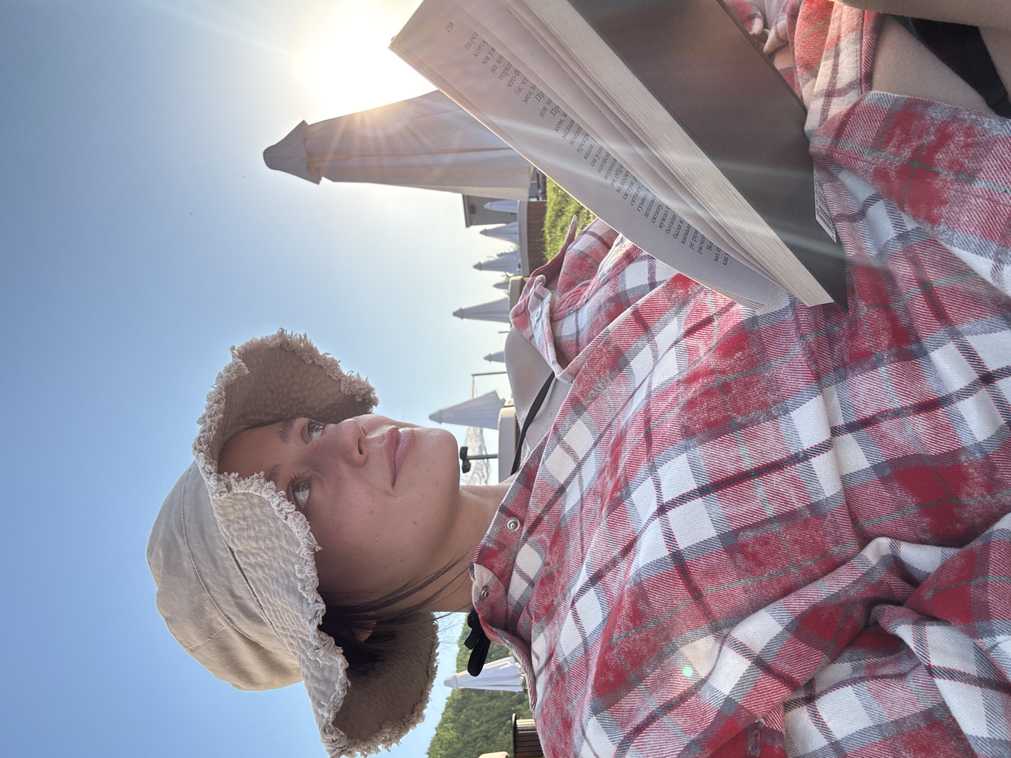 This image, captured in Дрокино, Russia, depicts a person relaxing outdoors on a clear, sunny day, seemingly immersed in a book. The photograph is taken from a low-angle perspective, looking up towards the subject, the expansive blue sky, and the bright sun, which creates visible lens flares.
The central subject is a young person, possibly female, attired in a red, white, and grey plaid flannel shirt and a light-colored bucket hat with frayed edges. She holds an open book, but her attention appears to be drawn upwards and away from the pages, suggesting a moment of contemplation, observation of the sky, or simply a pause in her reading. Her expression seems calm and thoughtful.
The setting is an outdoor leisure area, characterized by several white sun umbrellas visible in the background, hinting at a resort, beach, or patio environment. The strong, direct sunlight indicates it is likely late morning or early afternoon.
An open book is prominent in the frame, displaying page 258. The visible text, as identified by OCR, is in Russian and includes words such as "перег" (over), "котор" (which), "на ко" (on which), "что" (what), "мец хо" (German ho-), "янам" (to the fields/villagers), "узна" (found out), "хотно" (willingly), "вершен" (completed), "десять" (ten), "сятину" (hundreds), "гумно" (threshing floor), "дешево" (cheap), "нужно" (needed), "ботали" (worked), and "расчет" (calculation). These fragments suggest the book is prose, possibly a novel or historical account.
The image conveys a sense of peaceful relaxation and enjoyment of a pleasant, sun-drenched environment while reading.