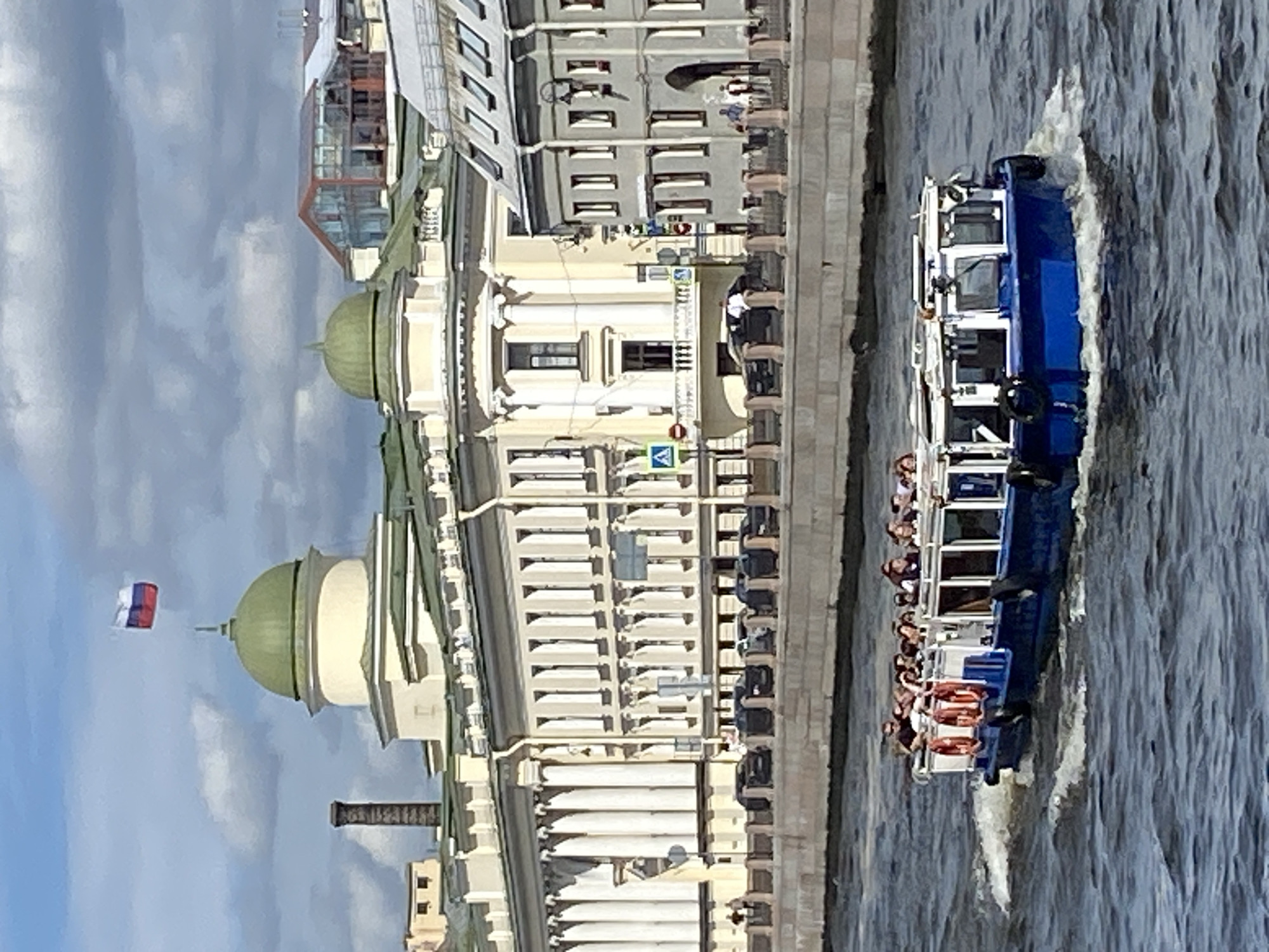 A tour boat with approximately twenty passengers is navigating a body of water, likely a canal or river, in Saint Petersburg, Russia. The boat is blue with white trim and has an enclosed cabin with windows. Several orange life preservers are visible on the side of the boat. In the background, a large, ornate building with a light yellow facade, white columns, and green domed roofs stands prominently. A Russian flag flies from a flagpole atop one of the domes. Further in the background, other buildings with a mix of architectural styles, including some with red-tiled roofs, are visible. A stone embankment lines the waterway in front of the buildings, with traffic lights and street signs also present. The sky is partly cloudy.