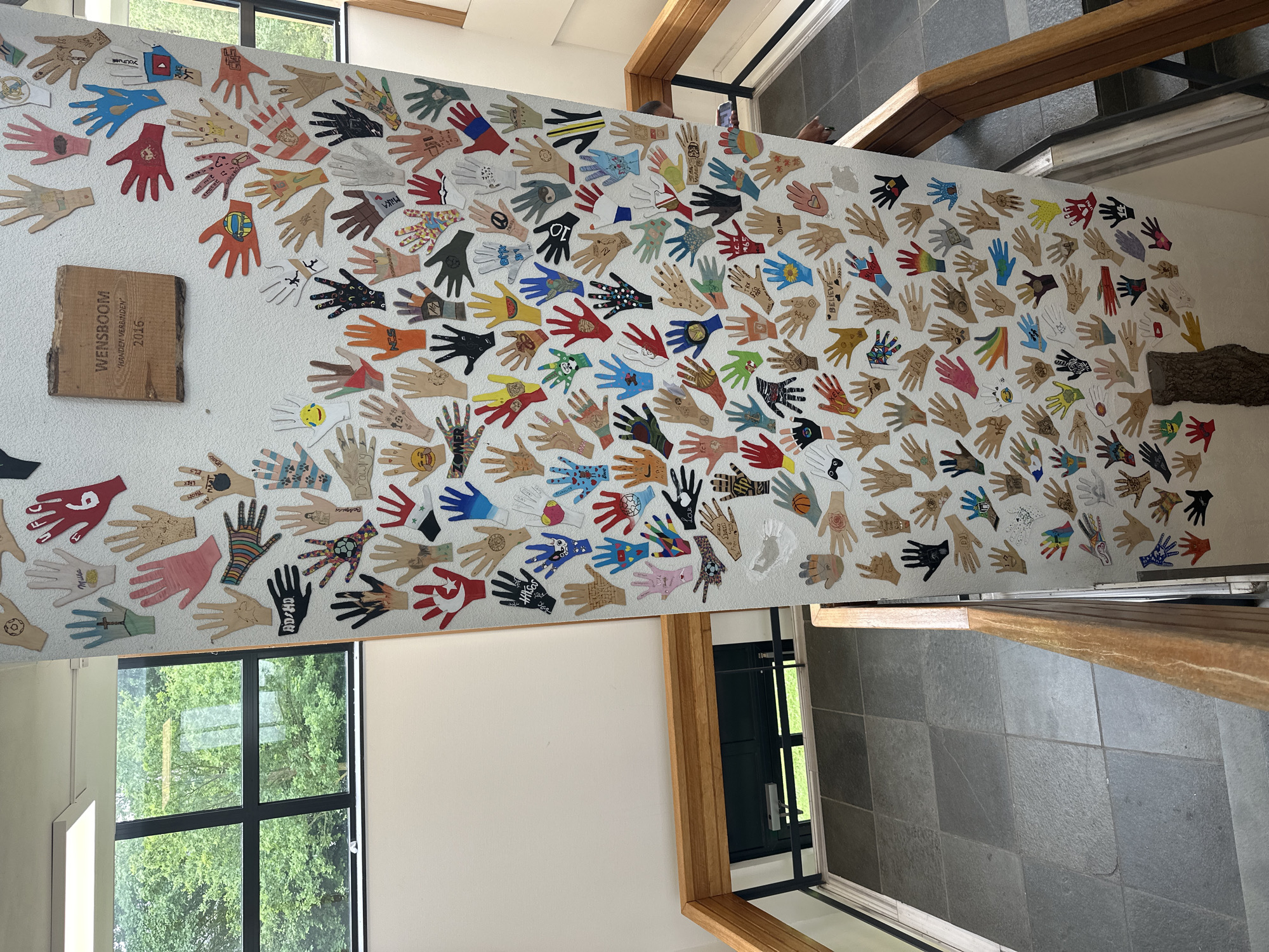 The image shows a colorful display of hand cutouts affixed to a white, curved wall that forms part of a staircase. The hand cutouts are diverse in color and design, with some featuring drawings, symbols, or text. A wooden sign with Dutch text "WENSBOOM HANDEN VERBINDEN 2016" is mounted on the wall above the hand cutouts. The staircase has wooden handrails and grey tiled flooring. In the background, through a large window, lush green trees are visible, suggesting an outdoor environment. The overall scene is likely from a school or community center in Borne, Netherlands, given the Dutch inscription on the sign. No people are clearly visible in the frame.