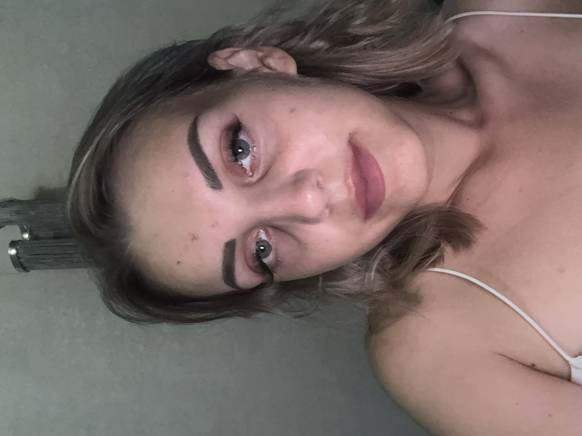 The image is a close-up, waist-up portrait of a young Caucasian woman with blonde hair. She is looking towards the camera with a neutral expression. She has visible under-eye bags and some blemishes on her forehead and cheek. Her makeup includes mascara and lipstick. She is wearing a white spaghetti strap top. The background is a plain, textured wall with some vertical elements visible at the top, possibly part of a light fixture or piece of furniture. There is no indication of the city of Yuzhno-Sakhalinsk, Russia, or any other specific location within the image.