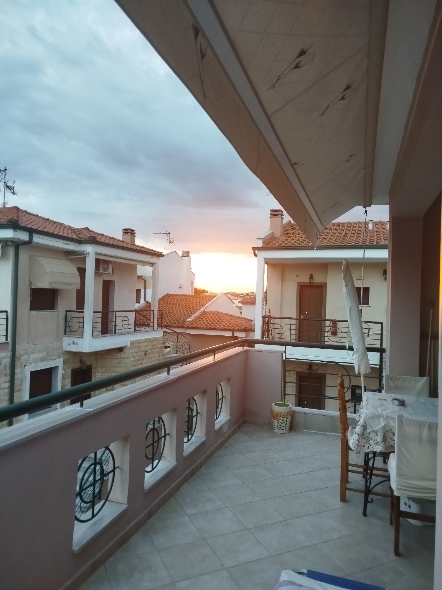The image depicts a view from a balcony overlooking a residential area in Sithonia Municipal Unit, Greece, during sunset. The balcony has a pink-colored railing with decorative circular metal grilles set within square openings. Beyond the balcony, several buildings with red-tiled roofs are visible. The sky is cloudy with the sun setting on the horizon, casting a warm glow on the scene. On the balcony floor, there are beige tiles, a white tablecloth draped over a table with chairs, a decorative pot, and a folded umbrella. In the foreground, a portion of an awning extends over the balcony. The overall scene suggests a peaceful evening atmosphere.