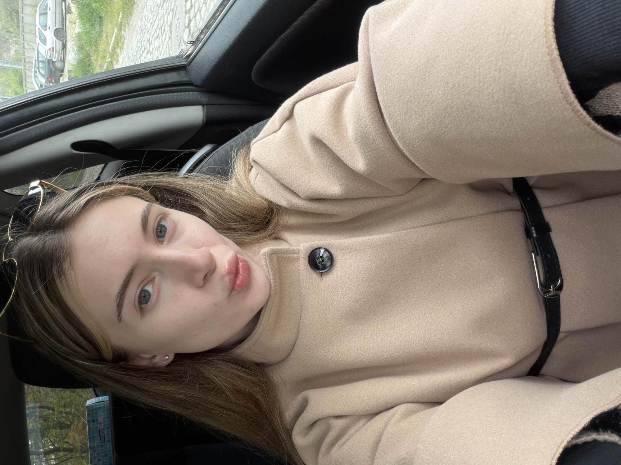 A selfie taken inside a car. The subject, a young woman with light skin and light brown hair with blonde highlights, is wearing a light beige coat with a black belt. She is looking at the camera with a slightly pouting expression.
The background shows the interior of a car. Through the car window, glimpses of an urban scene can be seen.
It is not possible to determine if the image was taken in the city of Plovdiv, Bulgaria.