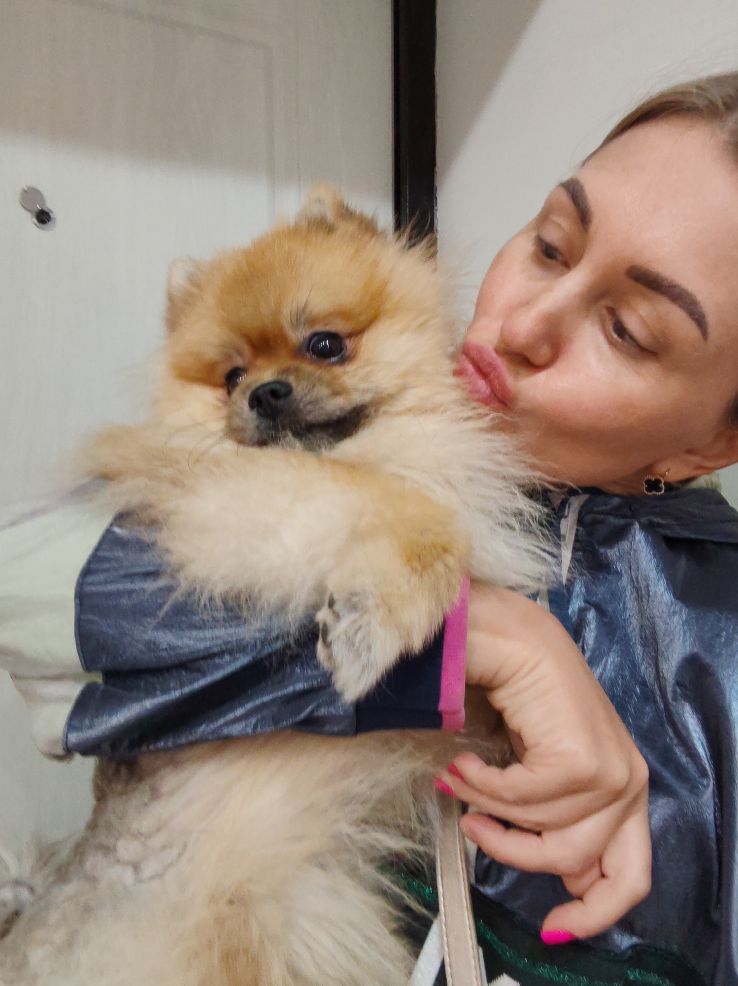 This is a close-up, indoor photograph of a woman holding a small, fluffy Pomeranian dog. The woman is leaning in to kiss the dog, with her lips puckered. She has blonde hair pulled back and is wearing a dark, shiny jacket with some lighter colored panels. Her fingernails are painted a bright pink. The dog is light brown and has dark eyes. The background appears to be a light-colored wall, possibly a door, with a small, circular object mounted on it. The lighting suggests it is daytime, but the indoor setting makes it impossible to determine the weather. The overall mood is affectionate and playful. There is no visible text in the image. The location context of Komsomolsk-on-Amur, Russia, does not provide any additional visual cues within the image itself.
