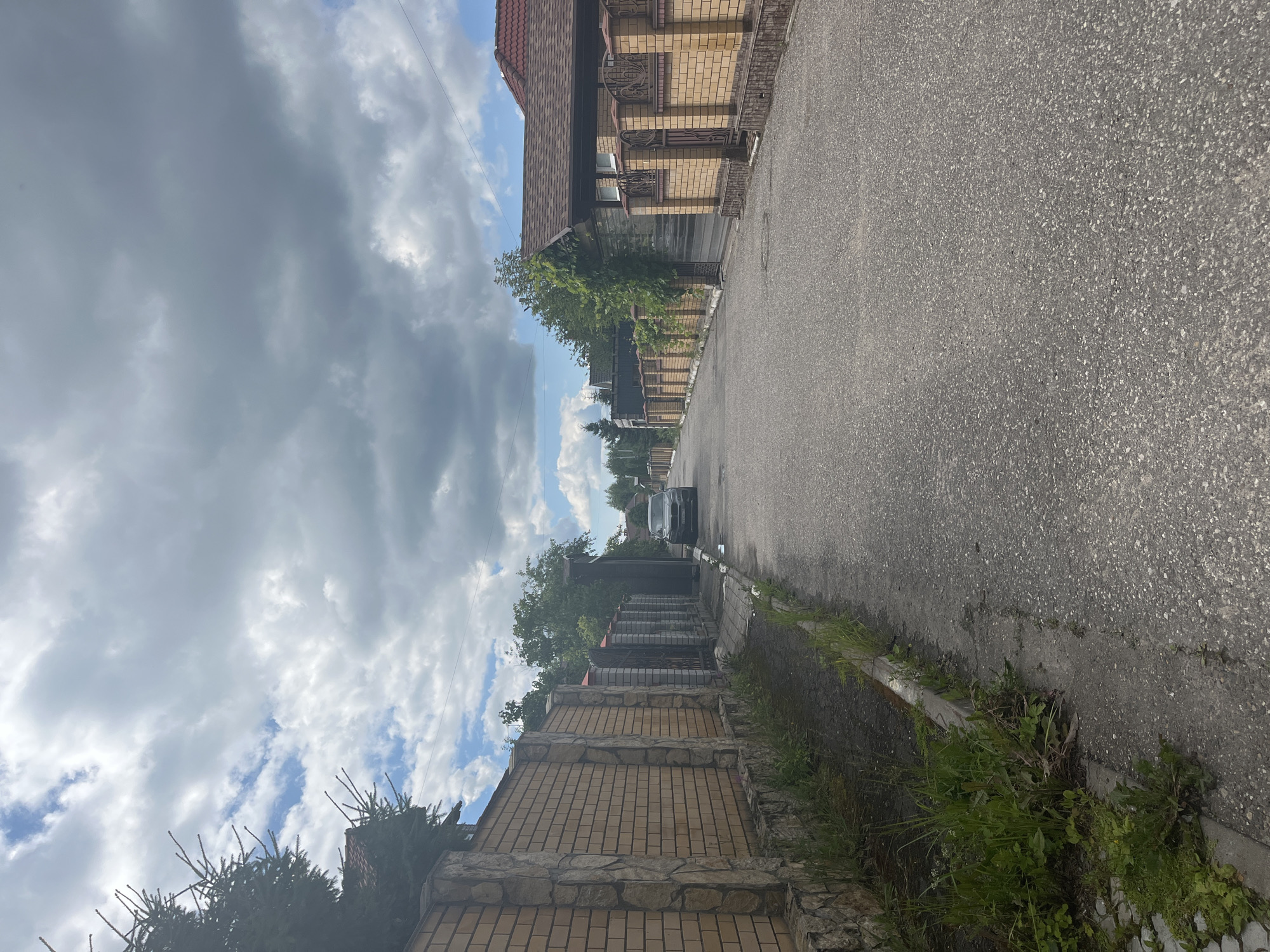 The image depicts a residential street in Orekhovka, Russia. A paved road runs through the center, flanked by brick fences and houses on either side. The houses have dark shingled roofs. There is a dark-colored car parked on the right side of the road. Vegetation, including grass and trees, is visible along the sides of the road and in yards. The sky is partly cloudy.