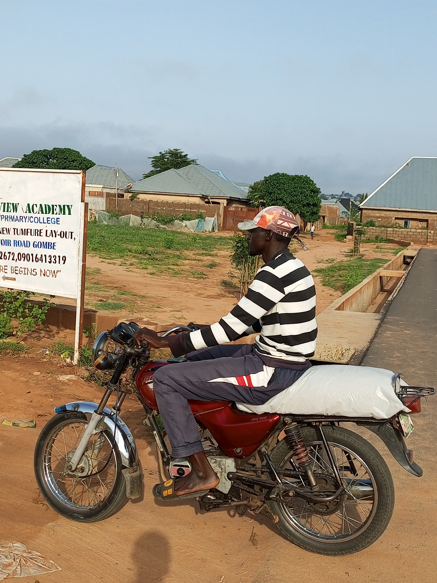A man wearing a black and white horizontally striped long-sleeved shirt, dark gray pants with a red and white stripe on the side, and a patterned baseball cap rides a red motorcycle on a dirt road. He is carrying a large white sack secured to the back of the motorcycle. To the left of the motorcycle is a white sign with green lettering. The sign reads "VIEW ACADEMY", "V/PRIMARY/COLLEGE", "NEW TUMFURE LAY-OUT,", "FOIR ROAD GOMBE", "2672,09016413319", and "RE BEGINS NOW" with an arrow pointing left. The background shows a few buildings with corrugated metal roofs, some vegetation, and a cloudy sky. The scene appears to be in Tempure, Nigeria.