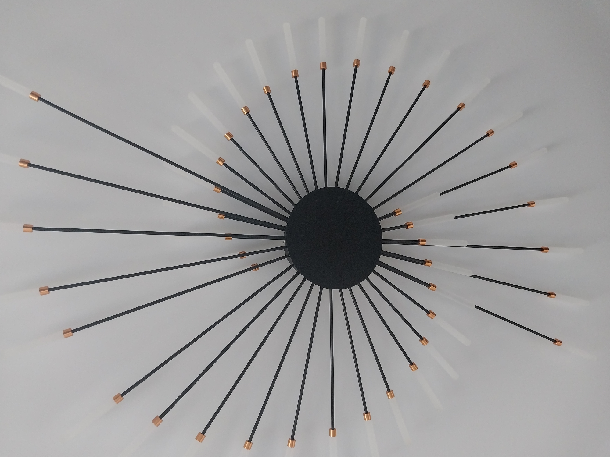 The image shows a modern sunburst-style wall clock with a black circular center. Thin black rods radiate outwards from the center, tipped with copper accents and clear or frosted glass elements. The clock is mounted on a plain white wall. The image appears to be taken from directly above. This object is located in Nizhneye Myachkovo, Russia.