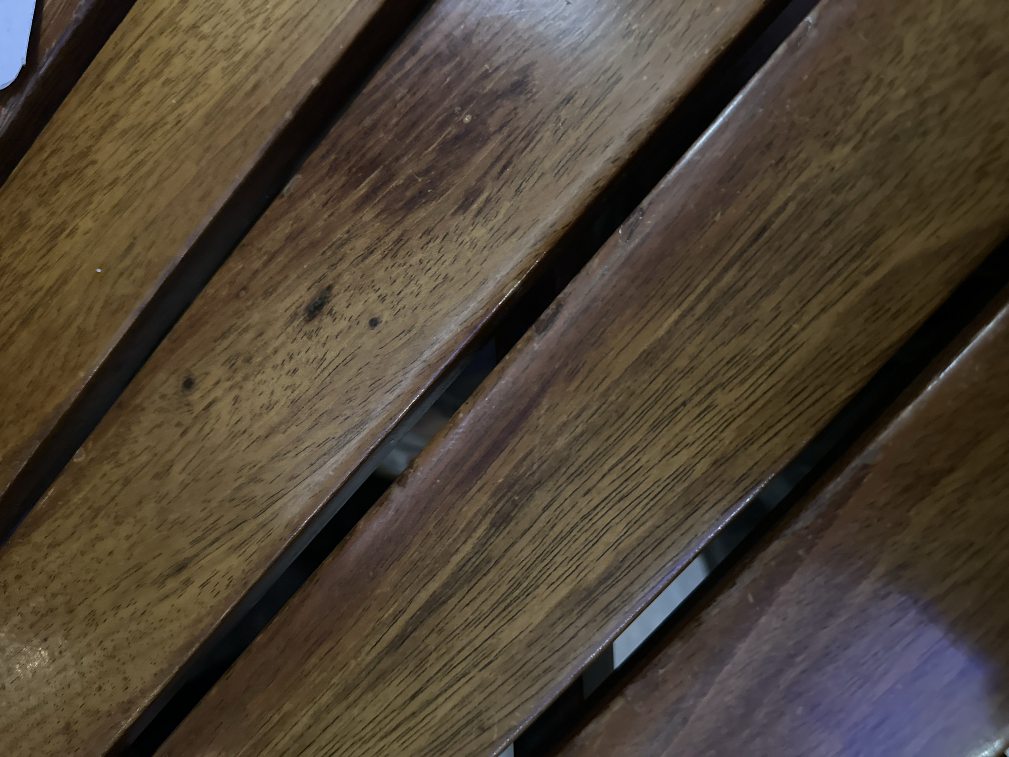 The image shows a close-up view of multiple parallel wooden slats. They are likely part of a bench or chair, displaying a varnished or polished finish. The wood grain is clearly visible. The location is unknown and there are no people visible in the image.