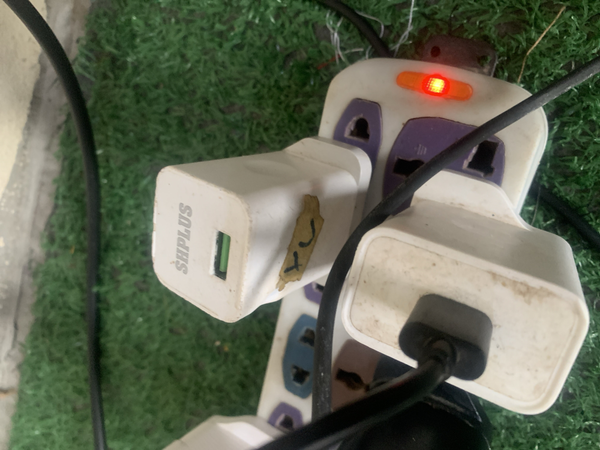 An overhead shot captures a close-up of a white power strip plugged into an unseen outlet, situated on a surface covered in artificial green turf. The power strip itself is a universal design, featuring multiple sockets with purple and blue accents. Two white USB charging adapters are plugged into the power strip. One adapter, in the foreground, has a black USB cable connected to it. Above this, another white adapter displays the embossed text "SHPLUS" and a green USB port. This adapter has a piece of yellow tape with a hand-drawn smiley face affixed to its side. Adjacent to the sockets, an orange illuminated switch glows red, indicating the power strip is active. The surrounding environment is dominated by the artificial turf, with a black cable running across the top of the frame and a hint of a concrete surface behind it. There are no people visible in the scene. This scene is not specifically located in Jauro Yinu, Nigeria.