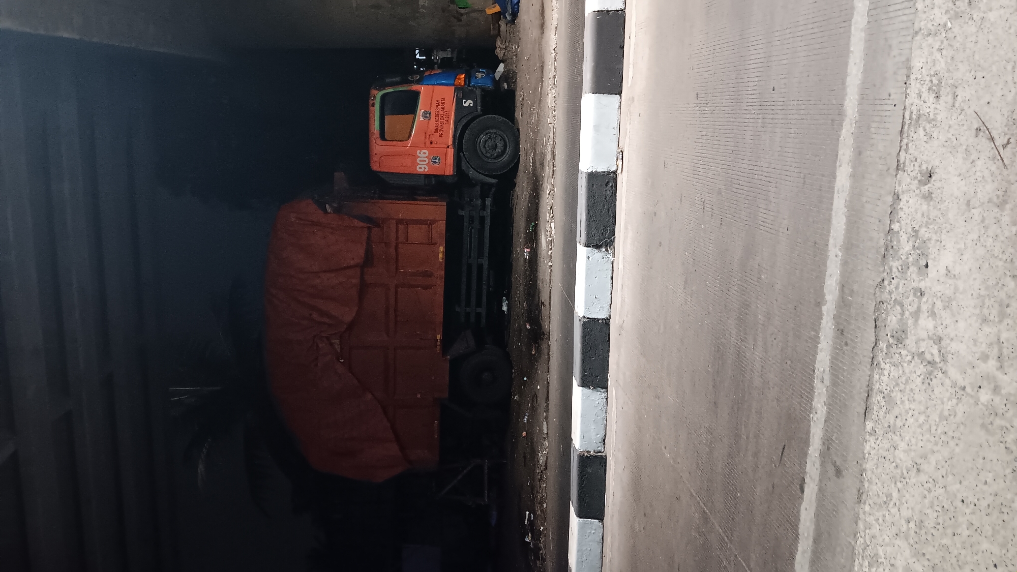 An orange and blue garbage truck with the number 906 prominently displayed on its side is parked under a concrete overpass. The truck's cargo bed is covered with a large orange tarp. The ground beneath and around the truck is littered with debris and trash. The scene appears to be in an urban environment, possibly a road or service area. The location is identified as South Jakarta, Indonesia, based on the text on the truck which reads "DINAS KEBERSIHAN PROVINSI DKI JAKARTA". The image is taken at night or in low light conditions, with the primary light source seemingly coming from the truck itself and artificial lighting from the overpass. A concrete barrier with alternating black and white painted sections runs across the foreground, separating the road from the area where the truck is parked.