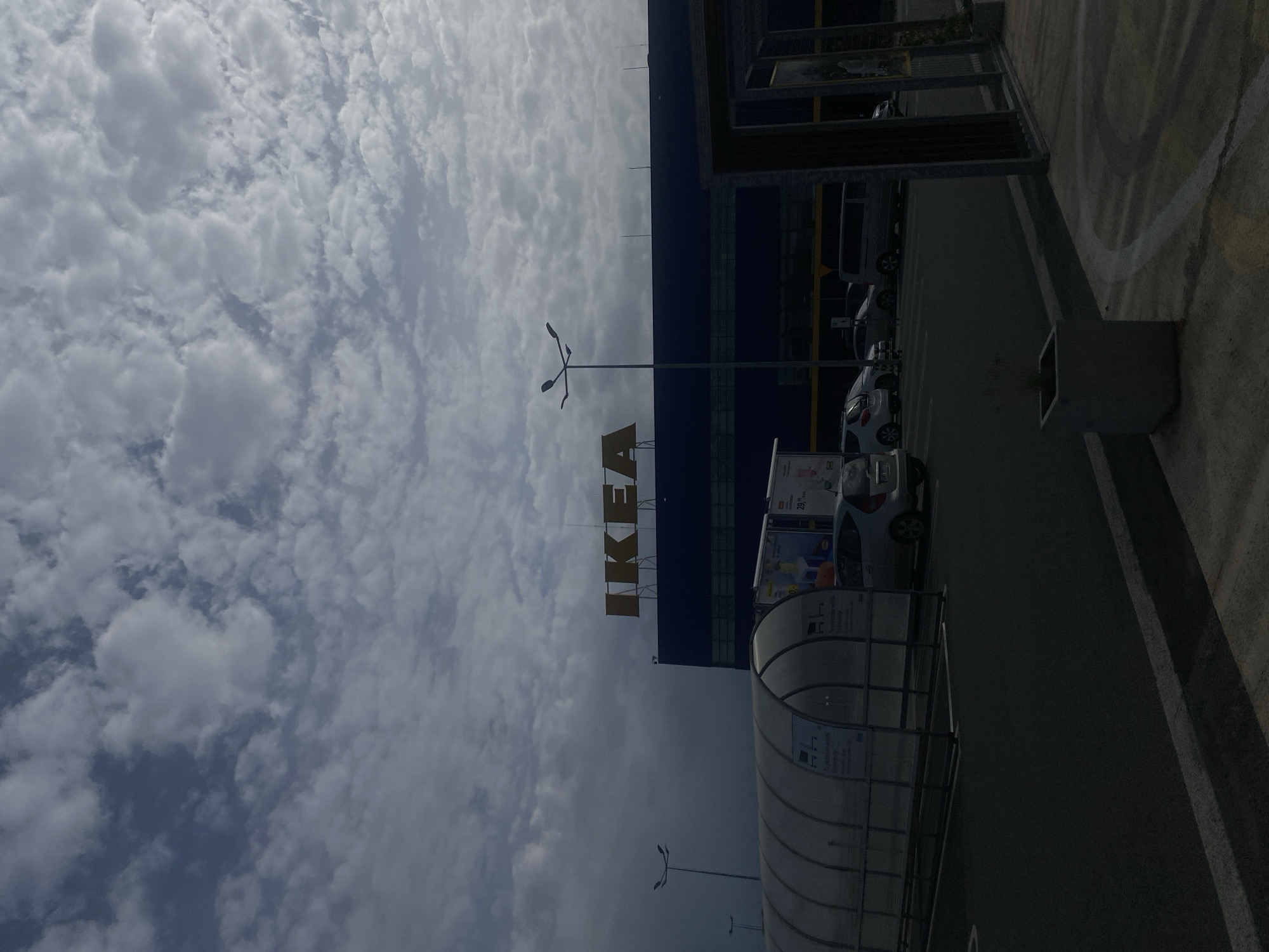 The image shows the IKEA store in Bielany Wrocławskie, Poland. The store is a large, blue building with the IKEA logo in large orange letters on the facade. In front of the store is a parking lot with several cars and a covered walkway for shopping carts. The sky is cloudy, with a mix of white and gray clouds. The lighting suggests it is daytime. The scene is presented in a static, observational manner.