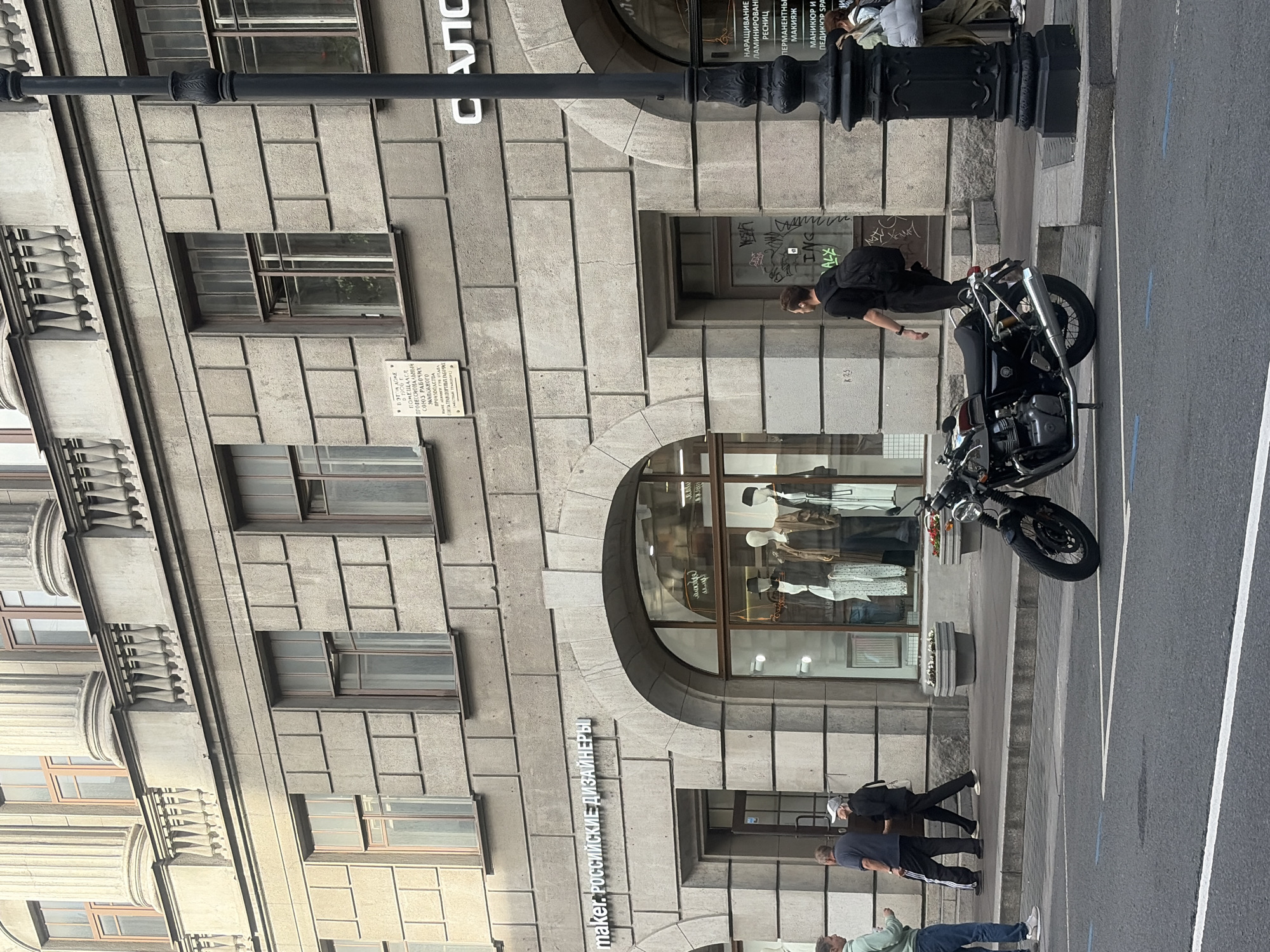 This image shows a street scene in Saint Petersburg, Russia. The main subjects are a motorcycle parked on the sidewalk and several people walking by. The motorcycle is black and appears to be a modern cruiser. One person, dressed in black and wearing a backpack, is leaning against the motorcycle. Further down the sidewalk, three other individuals are walking in the same direction. The building in the background is constructed of stone and features multiple windows, some with ornate detailing. A sign on the building reads "maker. РОССИЙСКИЕ ДИЗАЙНЕРЫ" which translates to "maker. RUSSIAN DESIGNERS." Another sign above a storefront window lists various beauty services like "НАРАЩИВАНИЕ РЕСНИЦ" (eyelash extensions) and "ПЕРМАНЕНТНЫЙ МАКИЯЖ" (permanent makeup). The scene is illuminated by daylight, suggesting it's daytime, and the weather appears to be overcast. The overall atmosphere is that of a typical urban street with a mix of commercial and residential architecture.