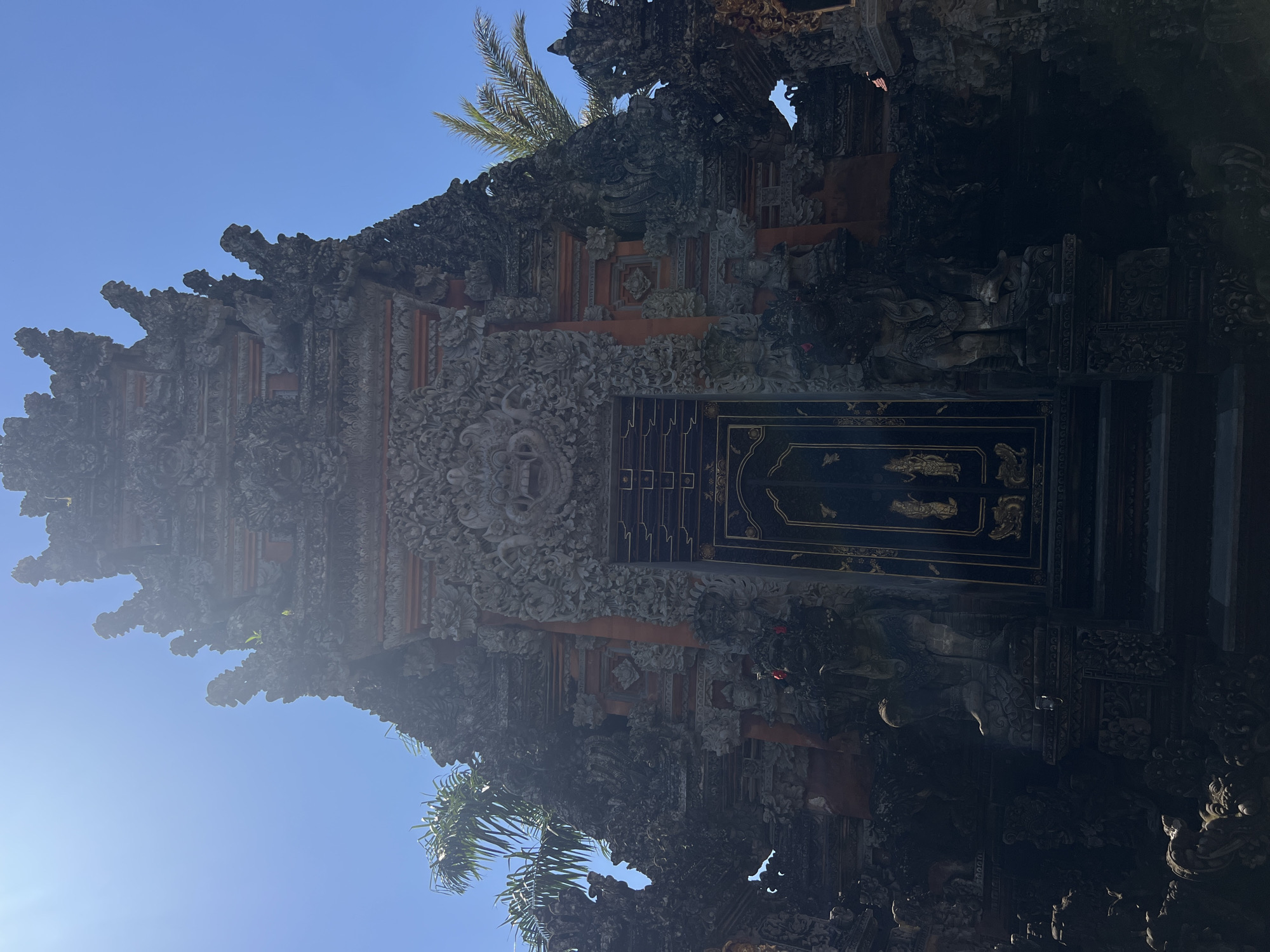 The image displays a detailed, ornate stone temple gate, characteristic of Balinese Hindu architecture, under a clear blue sky in the Ubud District, Indonesia. The gate, likely a Paduraksa or Candi Bentar, is predominantly dark grey and brown, heavily adorned with intricate relief carvings of deities, mythical creatures, and floral motifs. A prominent, menacing guardian face, resembling a Kala or Boma, is carved into the stonework directly above the dark, double-leafed main door. This door features golden-colored figures and patterns, appearing metallic against its dark background. Flanking the steps leading to the door are two large, stylized guardian statues (dwarapala), also carved from dark stone, standing guard. The upper sections of the gate exhibit multiple tiers of sculpted forms, some with small green sprouts growing from crevices. Sunlight from the upper left illuminates the sky and parts of the structure, creating a slight lens flare and casting deep shadows across much of the carved stonework, particularly on the lower half of the gate and the guardian statues. Sections of reddish-orange material, possibly brick or painted elements, are visible behind some of the carved stone. Palm tree fronds are visible on both the left and right sides, partially obscuring the background and indicating a tropical environment. No individuals or specific actions are depicted within the frame; the scene is static, focusing solely on the architectural subject.