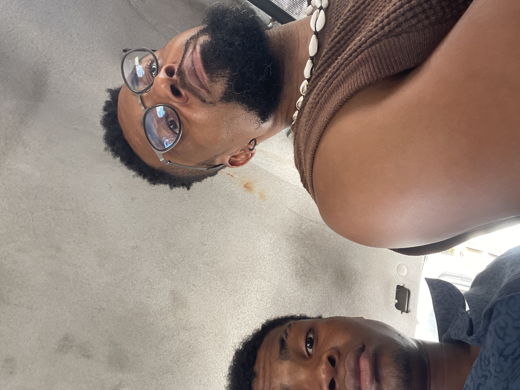 A rotated, low-angle shot captures two individuals with dark skin tones, positioned within what appears to be the interior of a vehicle, likely a car or bus, in Lagos, Nigeria. The camera is aimed upwards towards the ceiling, which is covered in a light gray, textured fabric, showing some light brown stains.
In the upper portion of the rotated image (appearing bottom right when viewed upright), the face and upper chest of one person are visible. They have dark, short curly hair and a beard, and are wearing round spectacles with thin metal frames. A black ring earring is visible in their left ear. They wear a brown ribbed or knitted sleeveless top, and a necklace of white cowrie shells rests on their chest. Their gaze is directed towards the camera, with a neutral expression. Reflections of ambient light sources are visible in their spectacle lenses.
In the lower portion of the rotated image (appearing top left when viewed upright), a second individual is partially visible, showing their face and shoulder. They have dark, short curly hair and a slight mustache, wearing what appears to be a dark, possibly patterned, collared shirt. Their expression is neutral, and they are looking slightly away from the direct camera angle. No visible text is present in the image.