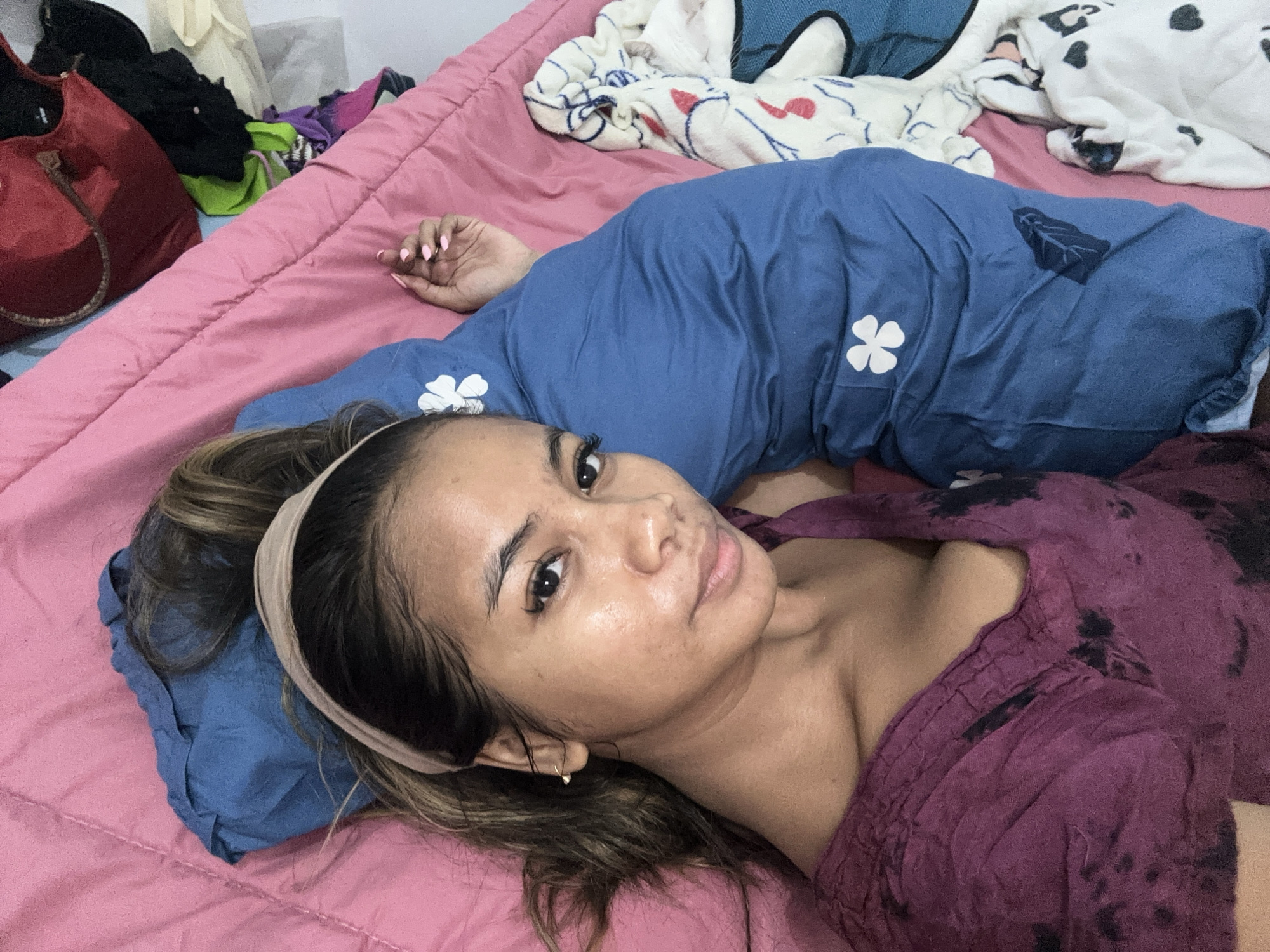 The image depicts a young woman lying on a pink bed in what appears to be a bedroom. She is propped up by a blue pillow with white flower patterns and is wearing a dark purple tie-dye shirt. Her head is resting on the pillow, and she is looking towards the camera. Her hair is dark brown and tied back with a beige headband. To the left of the bed, there is a pile of clothes and a red bag. The overall scene suggests a casual and relaxed setting, likely within a residential area in Sukawati, Indonesia.