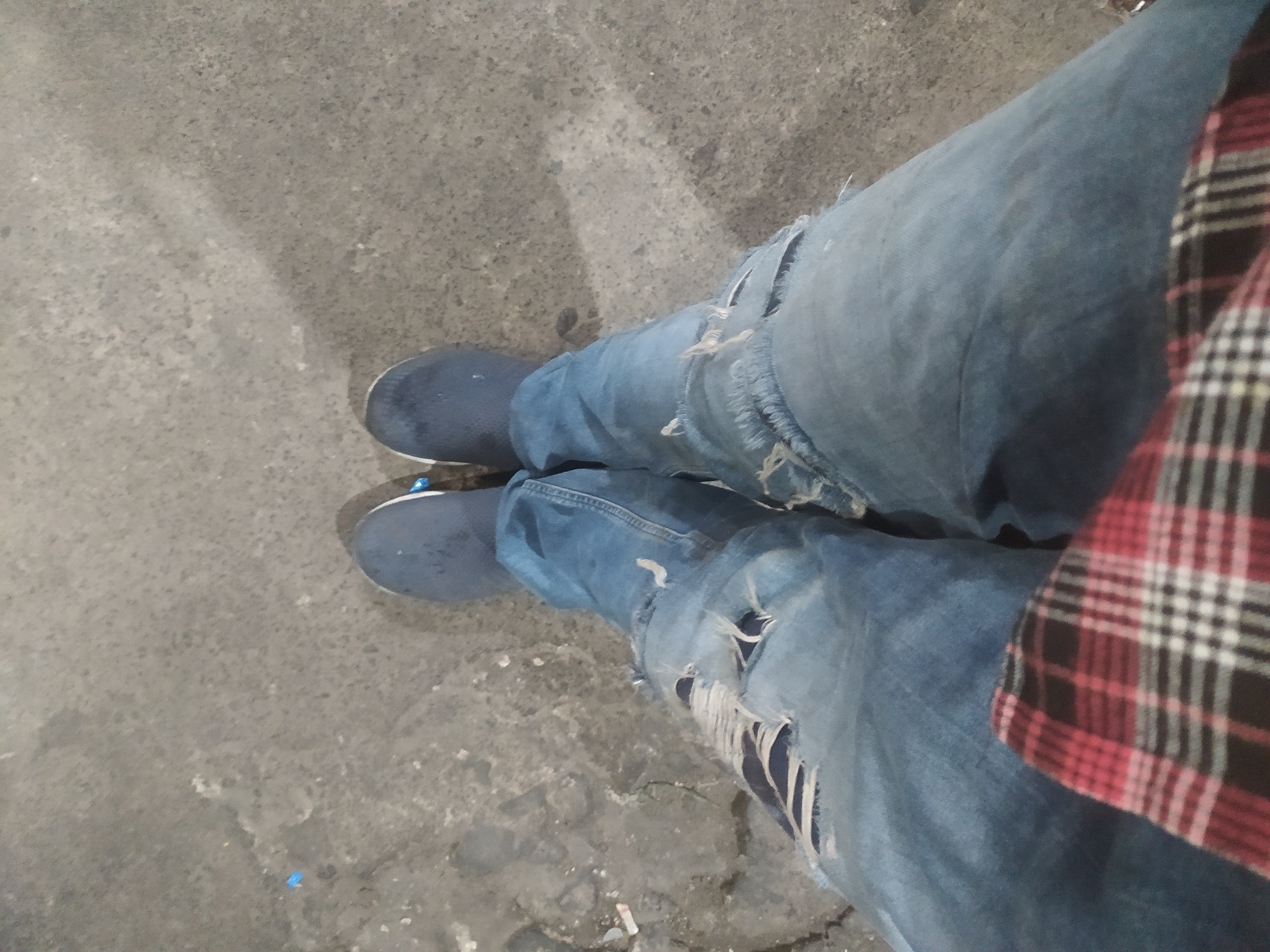 The image shows a close-up of a person's legs and feet, wearing ripped blue jeans and dark blue slip-on shoes. The person is standing on a grey, textured surface that appears to be asphalt or concrete, possibly a street or sidewalk. The lighting suggests it is daytime, with some shadows cast on the ground. A portion of a red, black, and white plaid shirt is visible in the foreground, draped over the person's lap. The overall impression is casual and urban.
The location is identified as Tuntang, Indonesia. Tuntang is a subdistrict in Semarang Regency, Central Java, Indonesia, known for its agricultural landscapes and proximity to the Tuntang River. While the image itself does not contain specific landmarks to pinpoint the exact spot within Tuntang, the scene is consistent with an outdoor urban or semi-urban environment found in Indonesia.