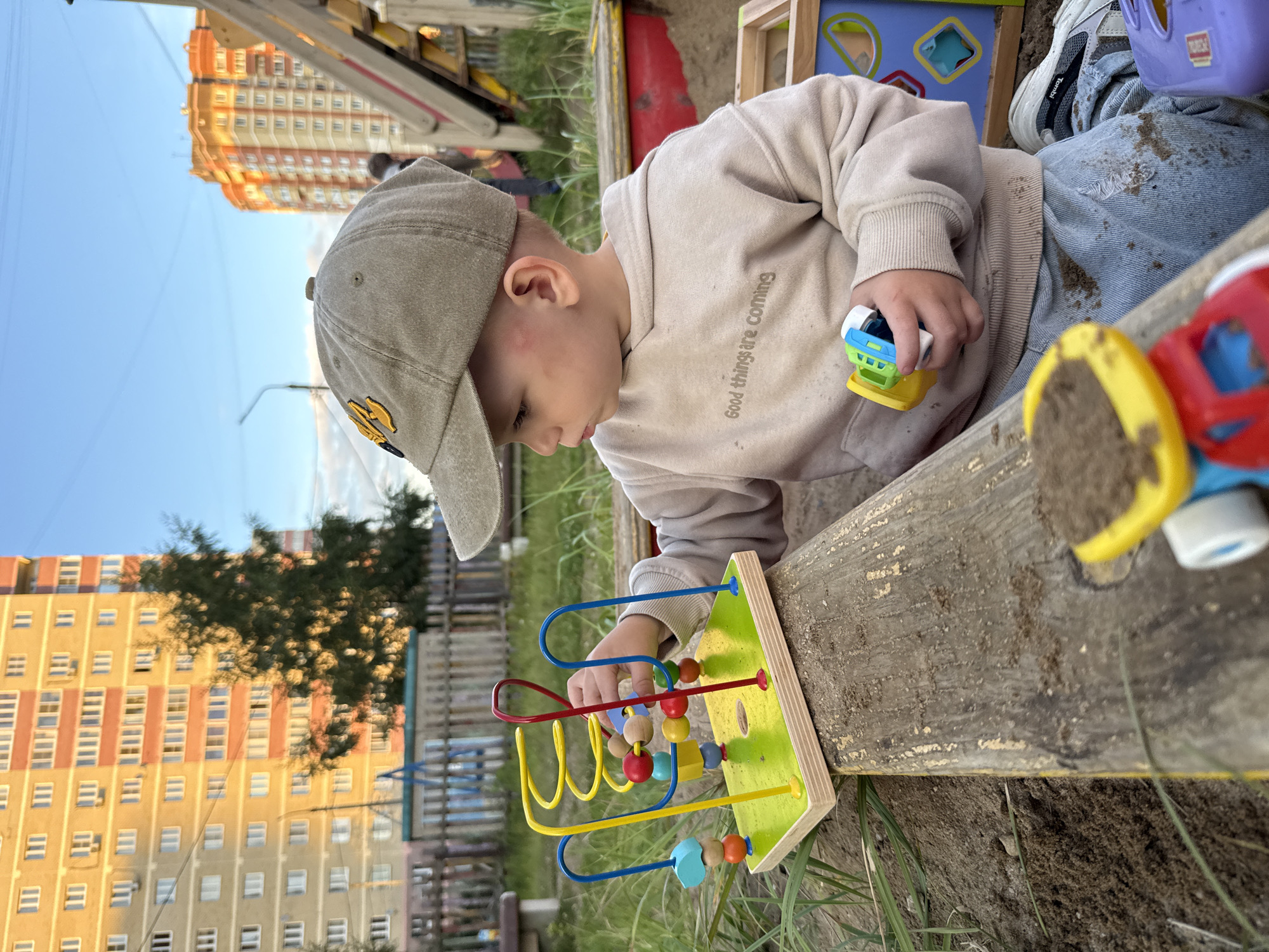 In a sandy playground in Rusanovo, Russia, a young child wearing a tan baseball cap and a beige hooded sweatshirt is playing with a wooden bead maze toy. The child is sitting in a sandbox, surrounded by sand and grass. In the background, two tall residential buildings are visible, one a lighter color with orange trim, and the other a darker orange. A green tree stands between the buildings, and a wooden fence encloses the playground area. The sky is a clear blue with a few faint clouds. The child is actively engaged with the toy, moving beads along the wire paths. Other toys, including a colorful toy truck, are scattered in the sand around the child.