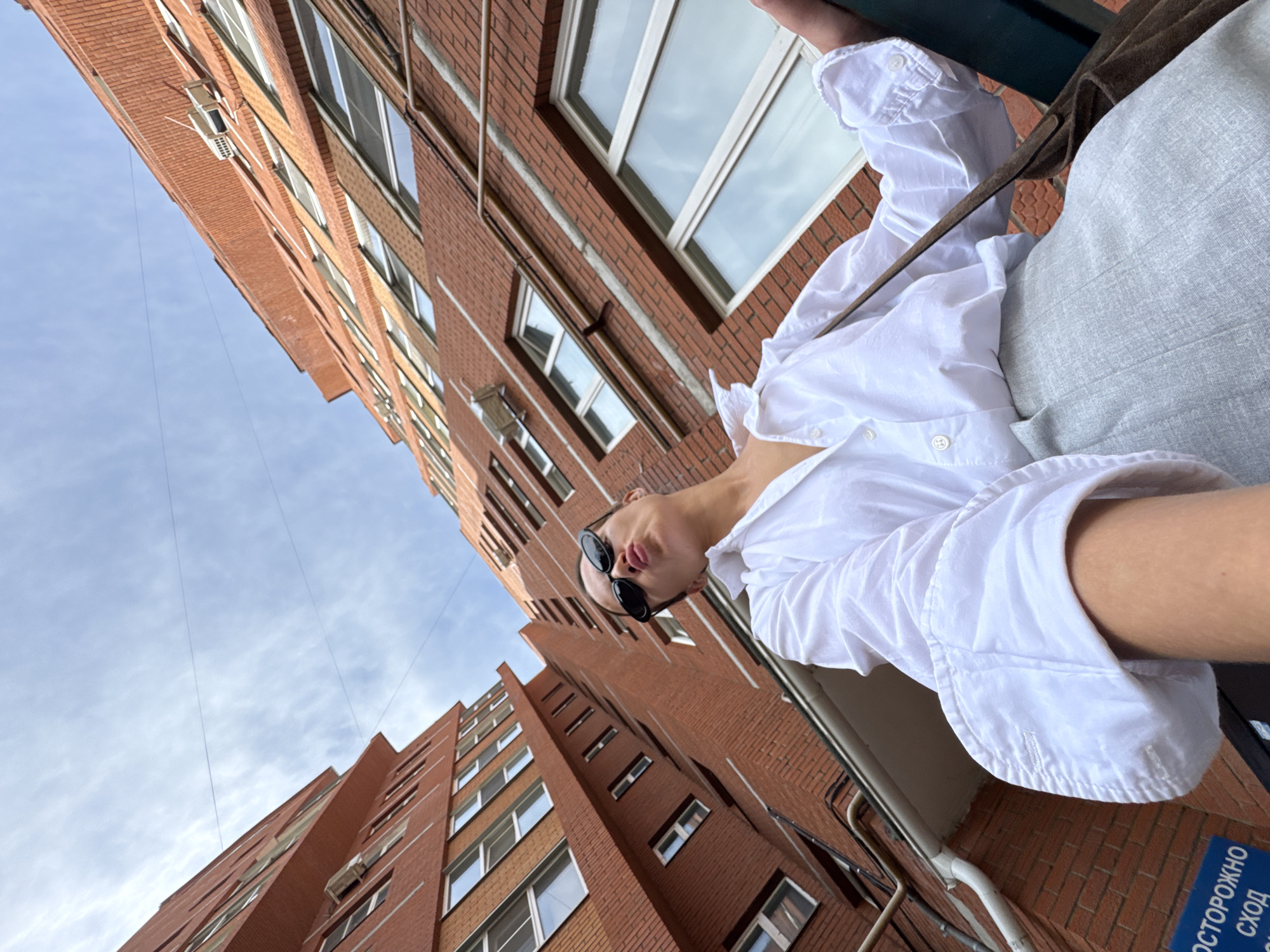 A woman is standing in front of brick buildings. She is wearing a white shirt and grey pants. She has sunglasses on and is puckering her lips. The buildings appear to be apartment buildings. The sky is visible with some clouds. There is a sign on the building that says "ОСТОРОЖНО СХОД". The location is the city of Chaikovsky, Russia.