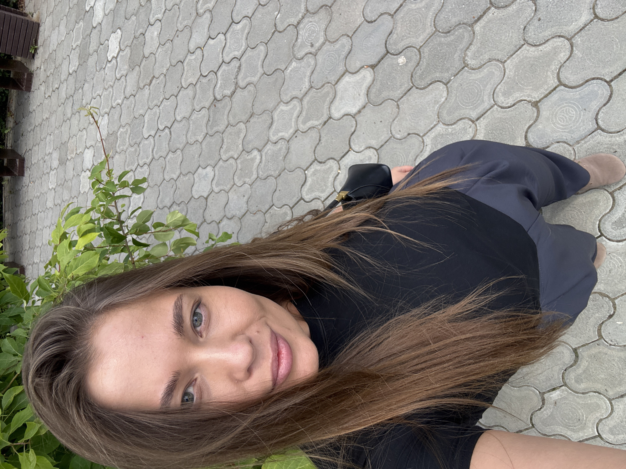 In a medium-high angle selfie, a woman with long, brown hair and light eyes is captured outdoors. She is wearing a black top and dark grey pants. The background consists of grey, interlocking pavers and some greenery. To the right of the frame, part of a dark wooden bench is visible, suggesting a park or public seating area. The scene is set in the city of Chernushka, Russia.