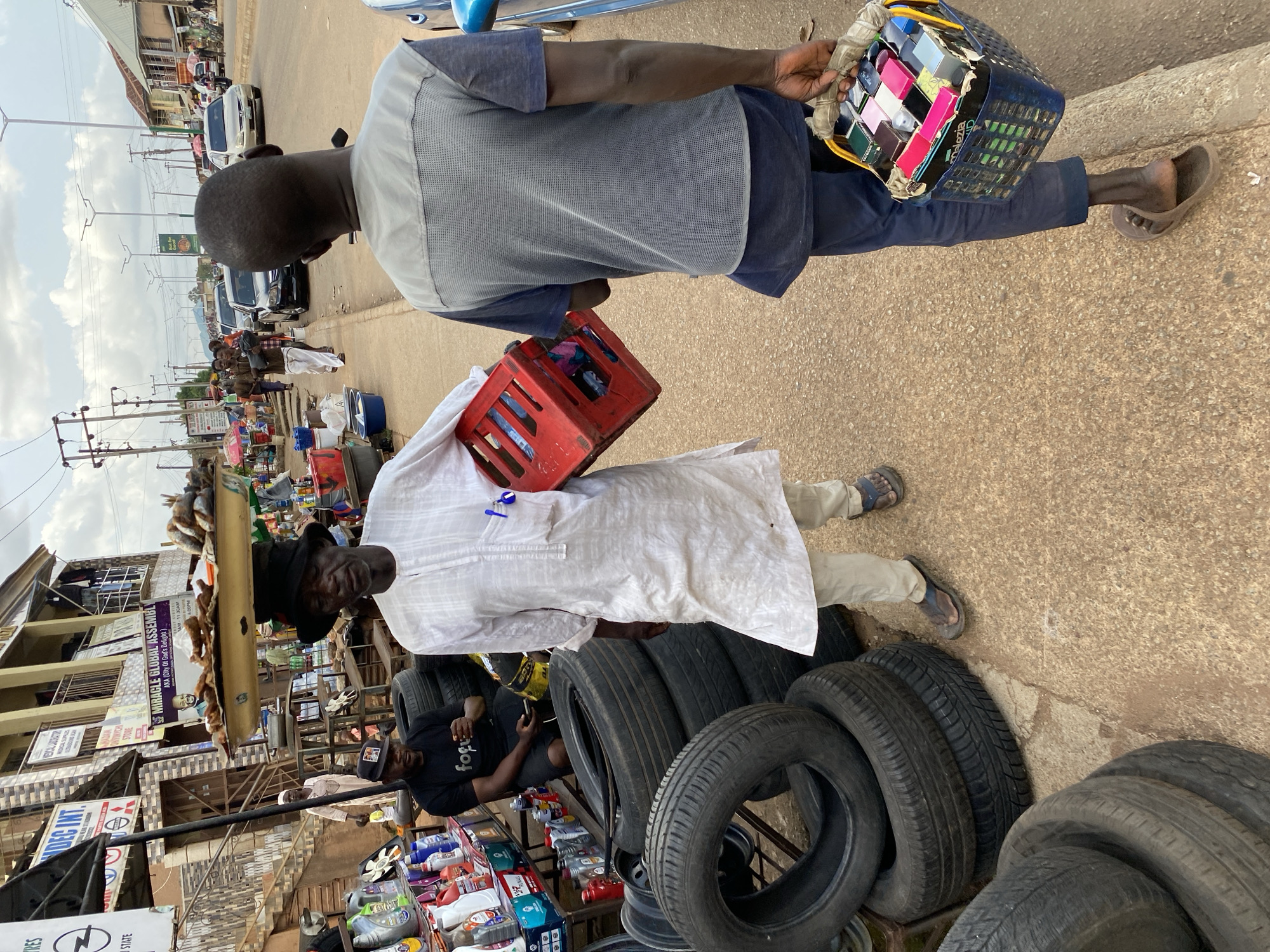 The image depicts a street scene in Bukuru, Nigeria. Two men are walking on a paved road. The first man carries a blue plastic basket filled with small, colorful boxes. He is wearing a grey and blue short-sleeved shirt and dark pants. The second man is holding a red crate. He wears a long white robe, beige pants, and dark sandals. Further down the street, a person in a black shirt is looking at a phone while sitting amidst a display of tires and various plastic bottles. Tires are also stacked nearby. Behind the tire display, a building is visible with multiple business signs displayed on its facade. Various other individuals and vehicles populate the background of the scene. Electrical wires stretch above the road.