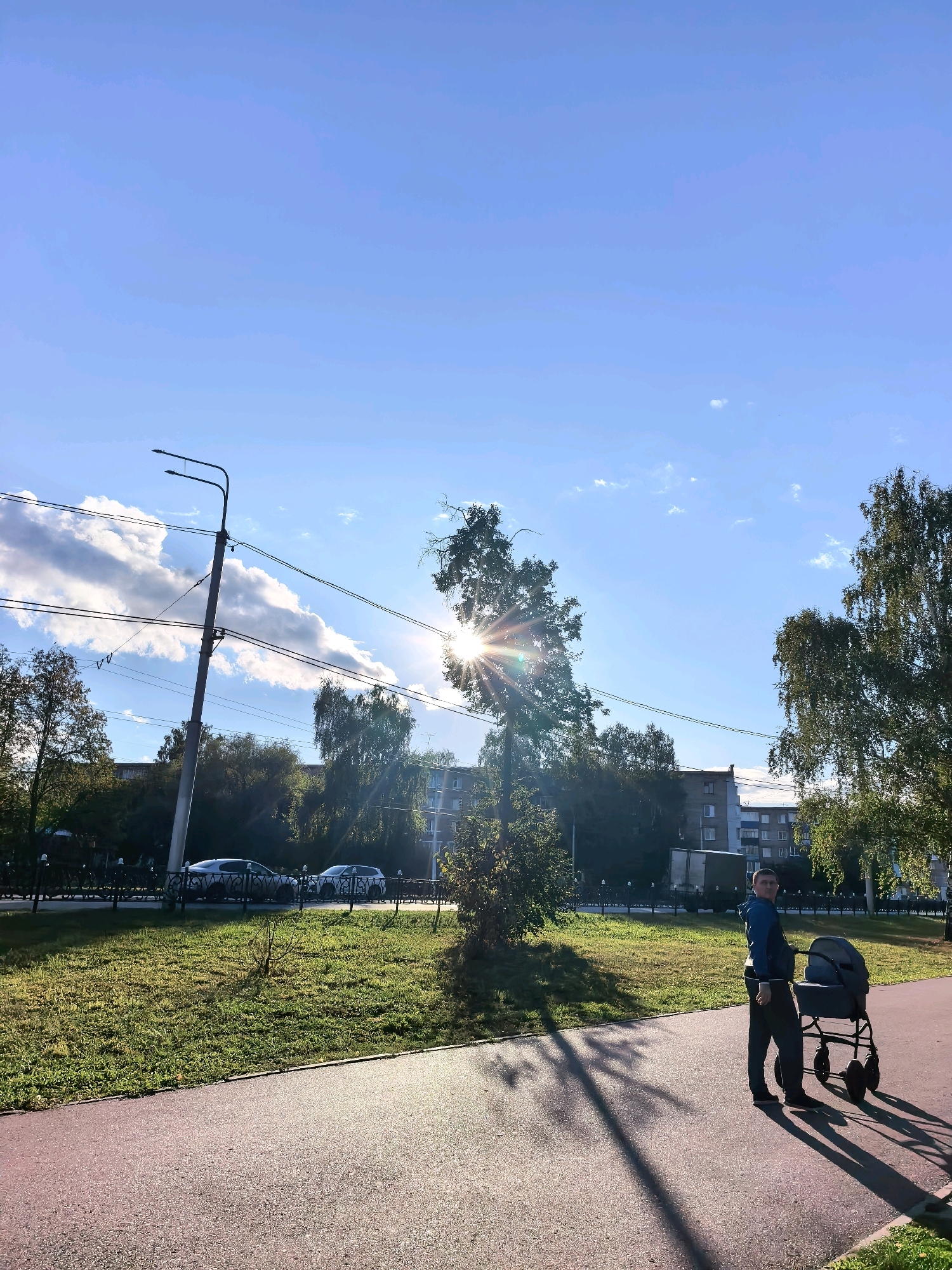 A man stands on a paved walkway in Sterlitamak, Russia, pushing a stroller. He is wearing a blue hooded jacket and dark pants. The sun is shining brightly through a tree, casting long shadows across the walkway. In the background, there are buildings, more trees, and cars parked along a street. The sky is a clear blue with a few white clouds. The walkway is bordered by a grassy area and a black metal fence.