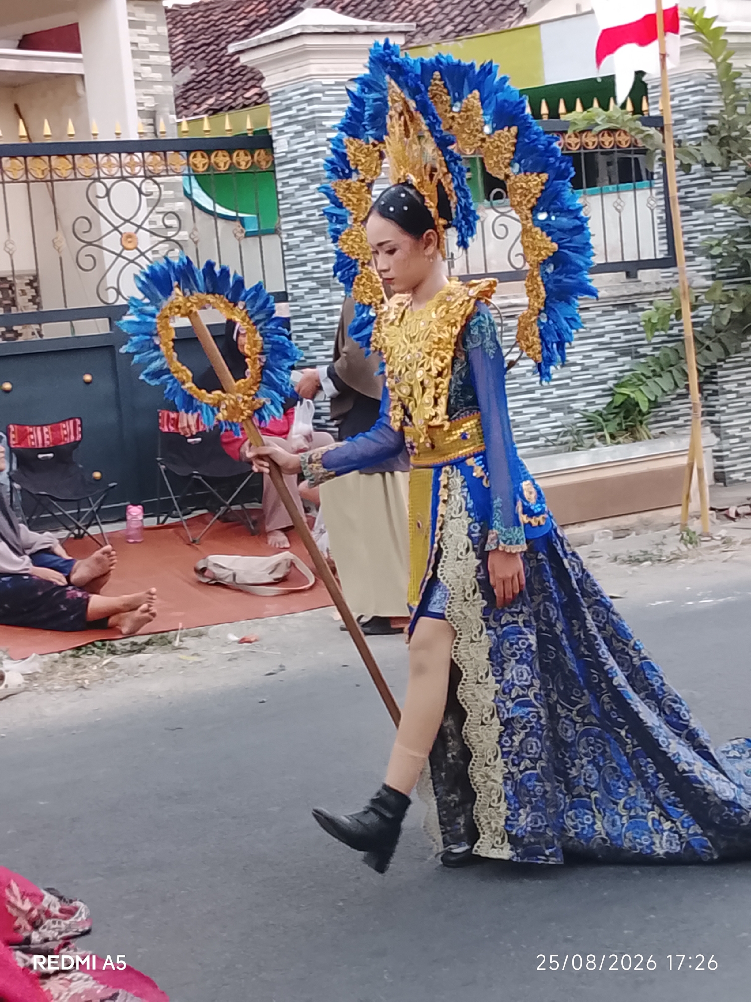 In Semanding, Indonesia, a person in a vibrant blue and gold costume with large, elaborate headwear is walking on a paved street. The costume features intricate gold embroidery and a long, flowing blue skirt with a patterned design. They are holding a wooden staff topped with a circular blue and gold ornament. In the background, there is a fence with decorative gold accents and a building with a tiled roof. Several people are seated on the ground to the left of the street, some wearing headscarves. The ground appears to be asphalt, and there is a small red mat on the side of the road. The scene suggests a parade or cultural event.