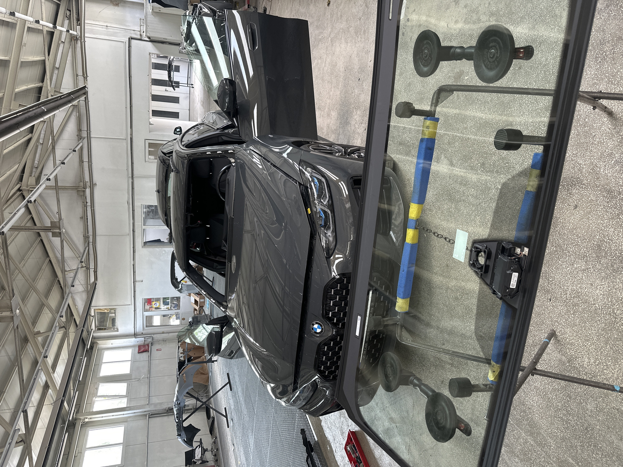 A dark grey BMW SUV is parked in an automotive repair workshop, appearing to be undergoing maintenance or repair. The vehicle's driver's and passenger's side doors are open, as is the trunk. Its front grille, prominently featuring the BMW logo, faces the viewer.
In the foreground, a removed windshield for the car is propped up against blue and yellow padded stands. Multiple black suction cups are attached to the windshield, and a black sensor module is affixed near its top center. The windshield's surface reflects the grey speckled workshop floor and overhead metal structures.
The workshop environment includes light-colored walls with multiple windows that provide natural lighting, augmented by artificial ceiling lights. Exposed metal beams and ventilation pipes run across the ceiling. In the background, another grey car bumper is visible on a stand, and a large white garage door can be seen. Various tools and equipment, including a red toolbox, are present around the workspace. Text reflected on the foreground windshield appears to show "BMW CAR" and "www.bimmer.org.ua". The scene depicts an active daytime automotive repair setting.