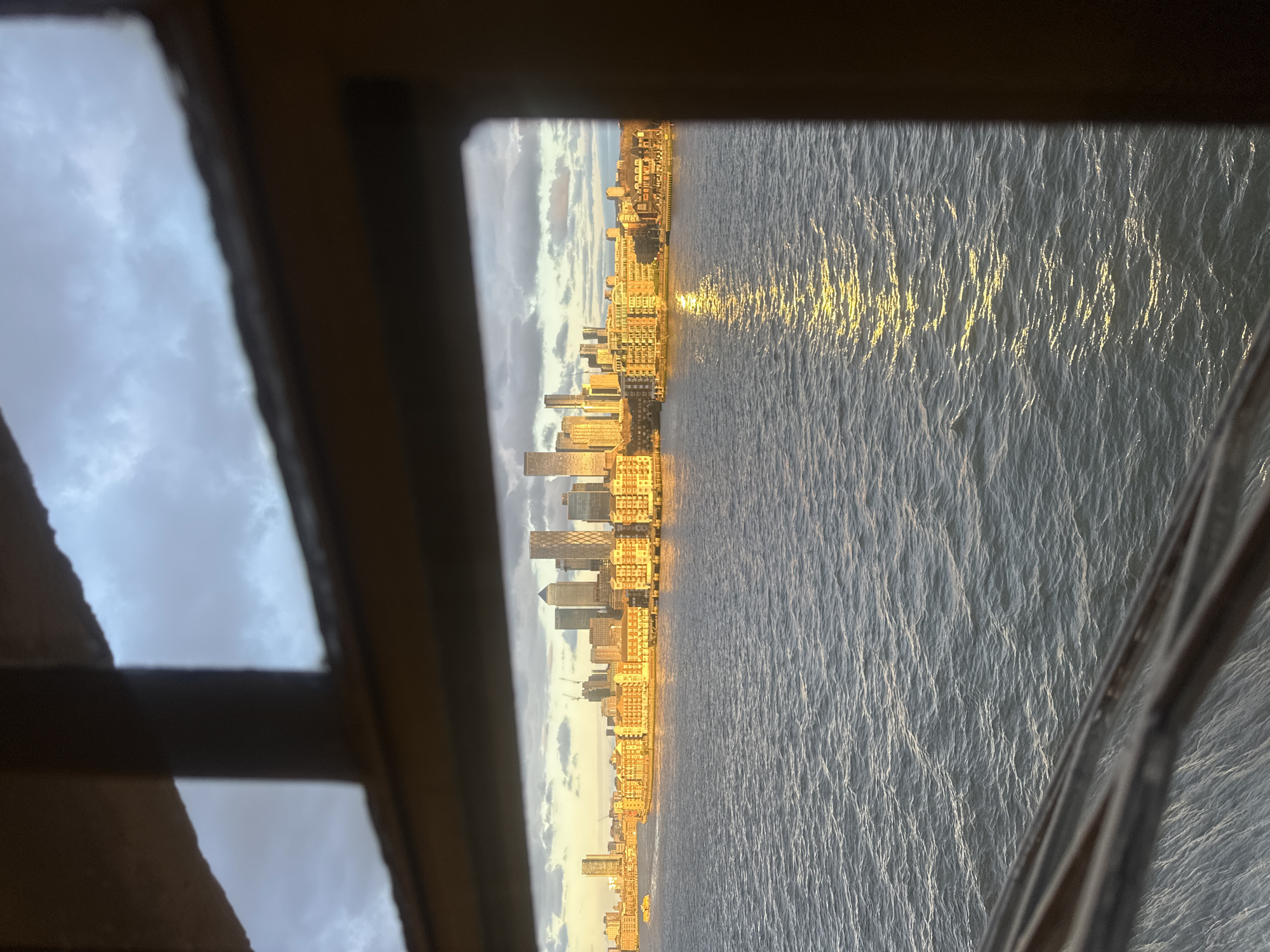 A view through a dark, multi-pane window frame depicts the River Thames with the Canary Wharf skyline in London, United Kingdom. The foreground presents choppy, dark blue-grey water, exhibiting a prominent golden reflection from low-angle sunlight, indicative of either sunrise or sunset. Across the river, the Canary Wharf financial district is visible, characterized by a cluster of modern skyscrapers and discernible construction cranes. A line of multi-story, light-colored residential or commercial buildings along the riverbank reflects the golden sunlight intensely. The sky above the cityscape is partially covered with grey and white clouds. A small, light-colored vessel is faintly visible on the water in the far-left distance. No individuals are clearly identifiable.