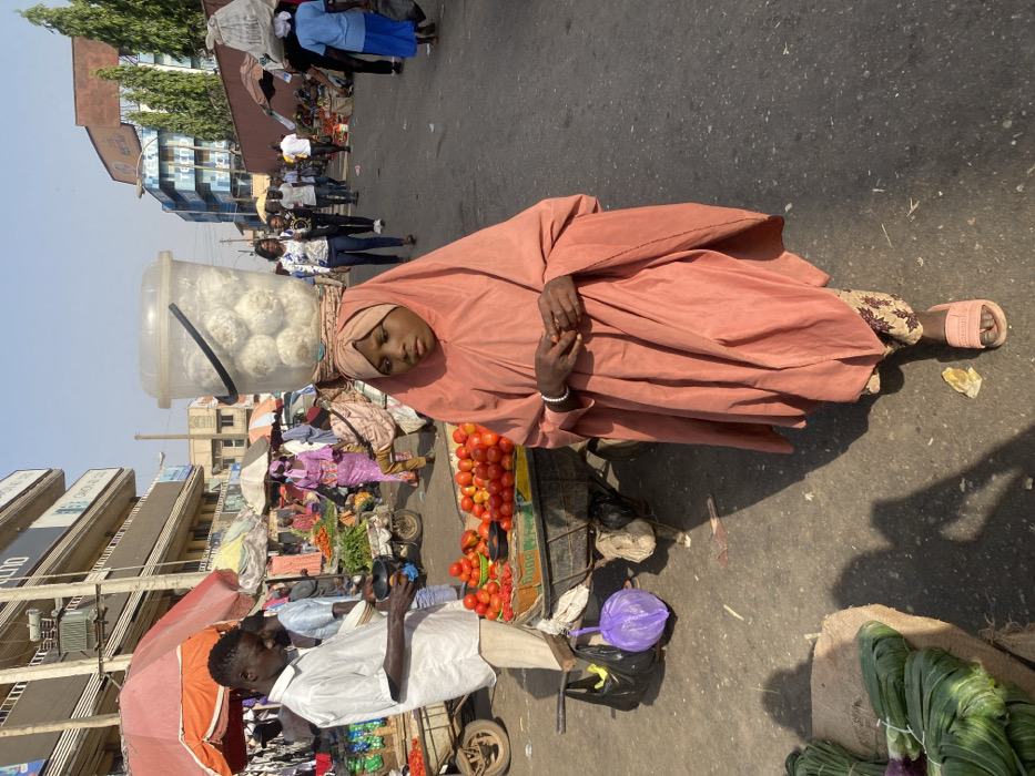 In a bustling outdoor market, a girl in a peach-colored hijab rests, a clear bucket filled with round, white food balanced on her head. She stands near a cart laden with tomatoes and other vegetables, near a man counting money. The surroundings suggest a vibrant, busy area with shops and pedestrians. The scene captures a moment of rest amidst the commerce of a typical market day.