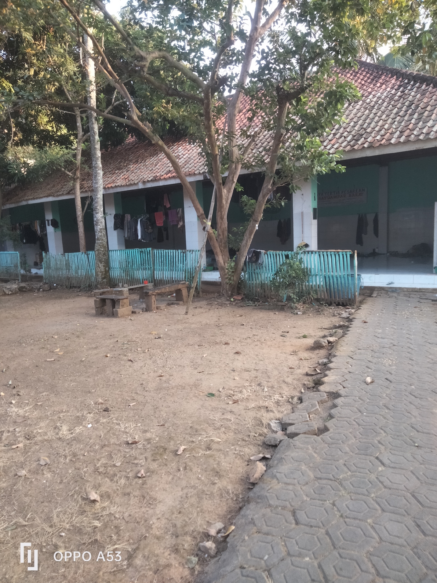 The image depicts an outdoor scene featuring a building with a tiled roof and a green-painted wall, possibly a public building or residence. Clothes are hanging on a line outside the building, suggesting everyday life. In front of the building, a turquoise wooden fence encloses a small area with some vegetation. To the left of the fence, there is a rustic wooden bench and table made of stone and wood, which appears to be a simple seating area. The ground in front of the building is unpaved, with dirt and scattered leaves. A paved pathway, composed of hexagonal concrete blocks, runs along the right side of the image. The overall impression is of a modest, perhaps rural or semi-urban setting, with elements of both a dwelling and a communal space. The presence of the "OPPO A53" watermark suggests the image was taken with a mobile phone. The location is identified as Badung, Indonesia, which is a regency in Bali known for its diverse landscapes, including beaches and cultural sites. This particular scene does not immediately convey a specific landmark or iconic feature of Badung, but rather a slice of local life.