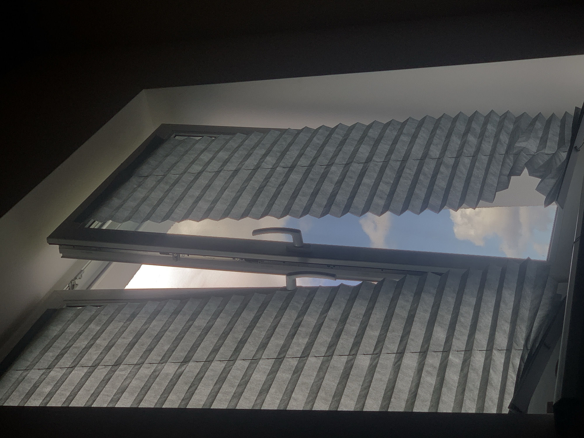 The image shows a partially opened window with pleated blinds. The blinds on the left are mostly intact but pulled down, while the blinds on the right are significantly damaged and torn, with large sections missing. Through the open window, a bright sky with clouds is visible. A white window frame with a silver handle is present. The interior appears dimly lit, contrasting with the bright exterior. The scene is shot from a low angle, looking upwards towards the window. The image does not provide any specific visual cues to indicate its location in Minsk, Belarus, or any other city.