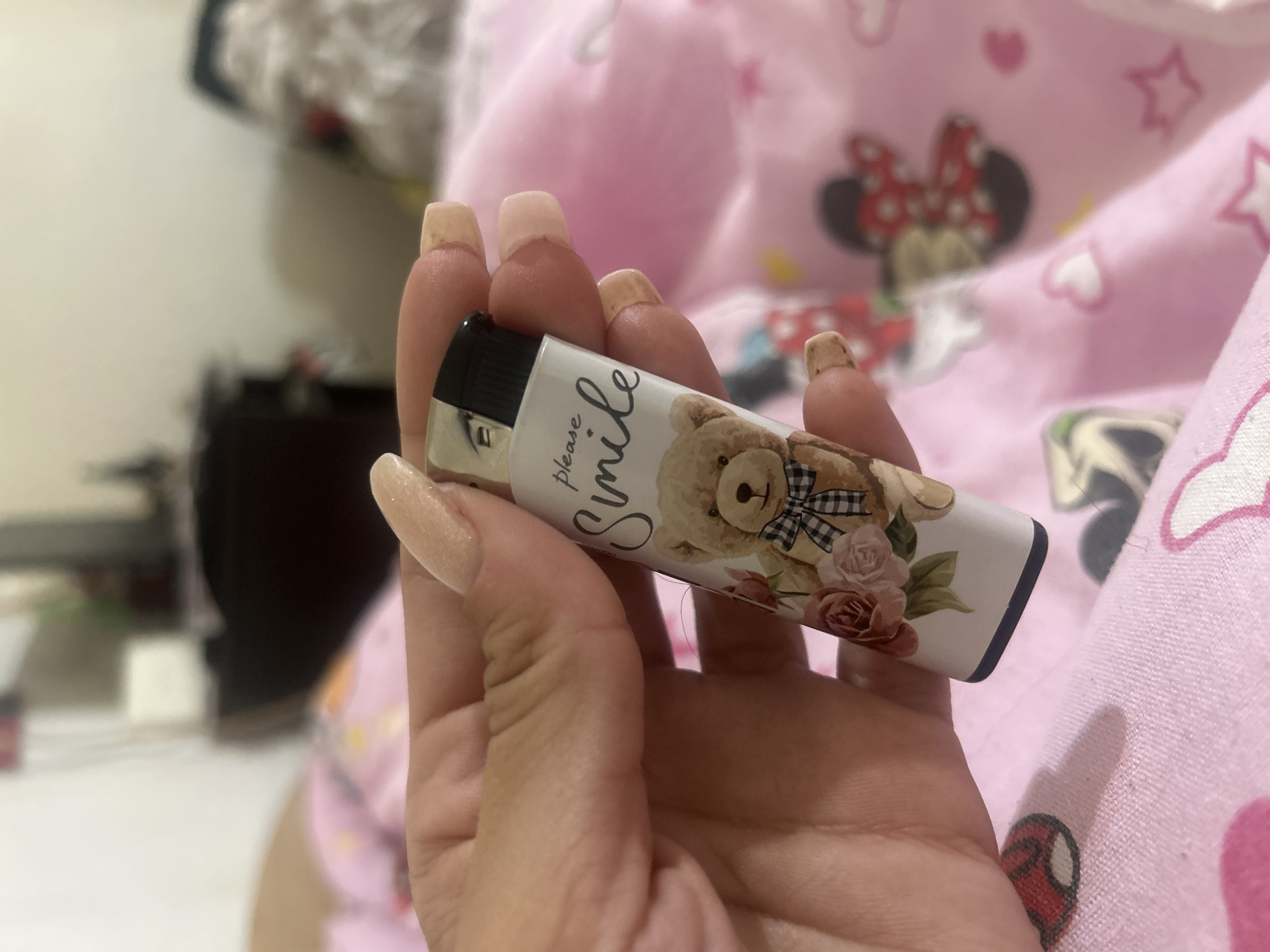 A person's hand, with long, light-colored, manicured fingernails, holds a white lighter decorated with a cartoon teddy bear, roses, and the words "please smile." The lighter has a black top. The background is blurred, but appears to be a pink fabric with Minnie Mouse designs, suggesting an indoor setting, possibly a bedroom. The location in Perushtitsa, Bulgaria, is not discernible from the image.