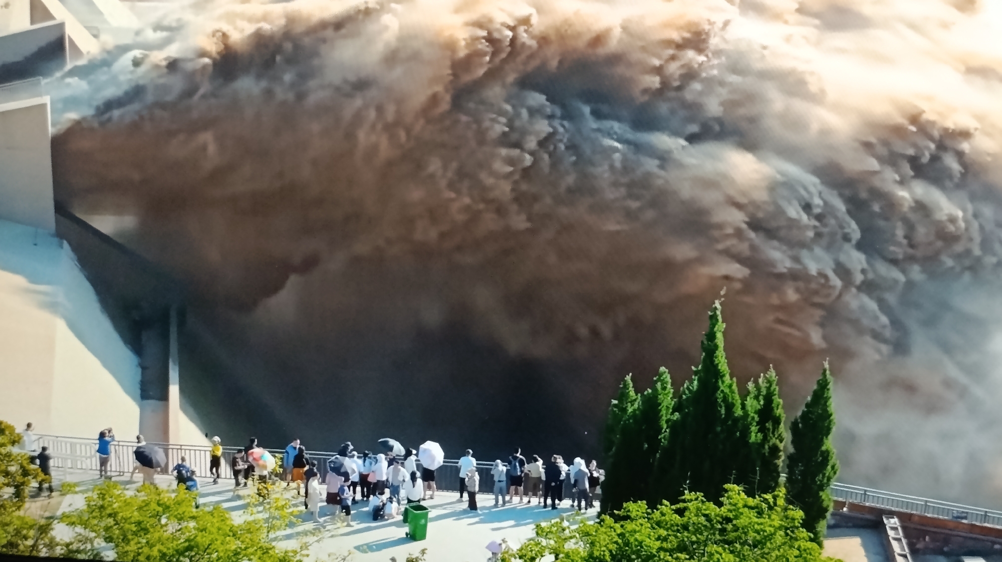 The image shows a powerful surge of muddy water being released from a large concrete structure, likely a dam or spillway. The water creates a massive, turbulent wave that dominates the background. In the foreground, a group of people are gathered on a walkway, observing the spectacle. Some are holding umbrellas, possibly for shade or protection from spray. Several individuals are taking photographs with their phones. Lush green trees are visible in the lower foreground and to the right, contrasting with the raw power of the water. The scene appears to be an organized viewing area, with a railing separating the onlookers from the drop. The location is identified as the Dubăsari town council in Moldova.