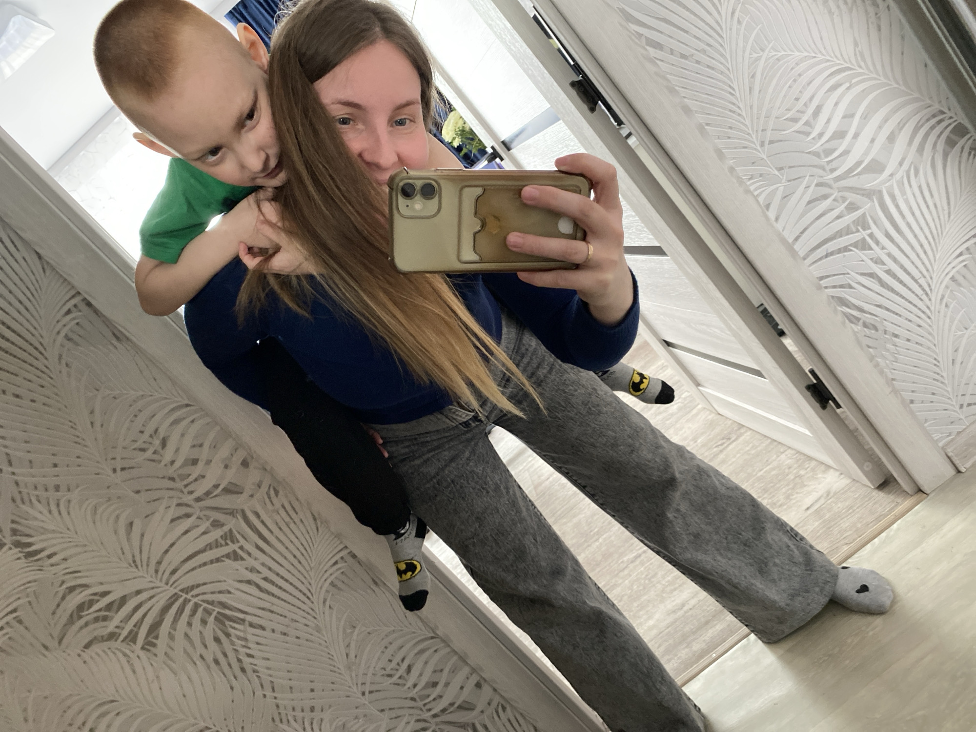 A woman and child are taking a selfie in an interior location. The woman is holding a smartphone with a protective case, facing the camera, while giving a piggyback ride to a young boy wearing a green shirt. The woman has long brown hair and is wearing a dark blue sweater and gray jeans. The boy is wearing gray socks with a batman logo. The location includes a white door, wall with a palm leaf print, and wood floor. It's impossible to verify the precise location in Noviy, Russia, from the given image.